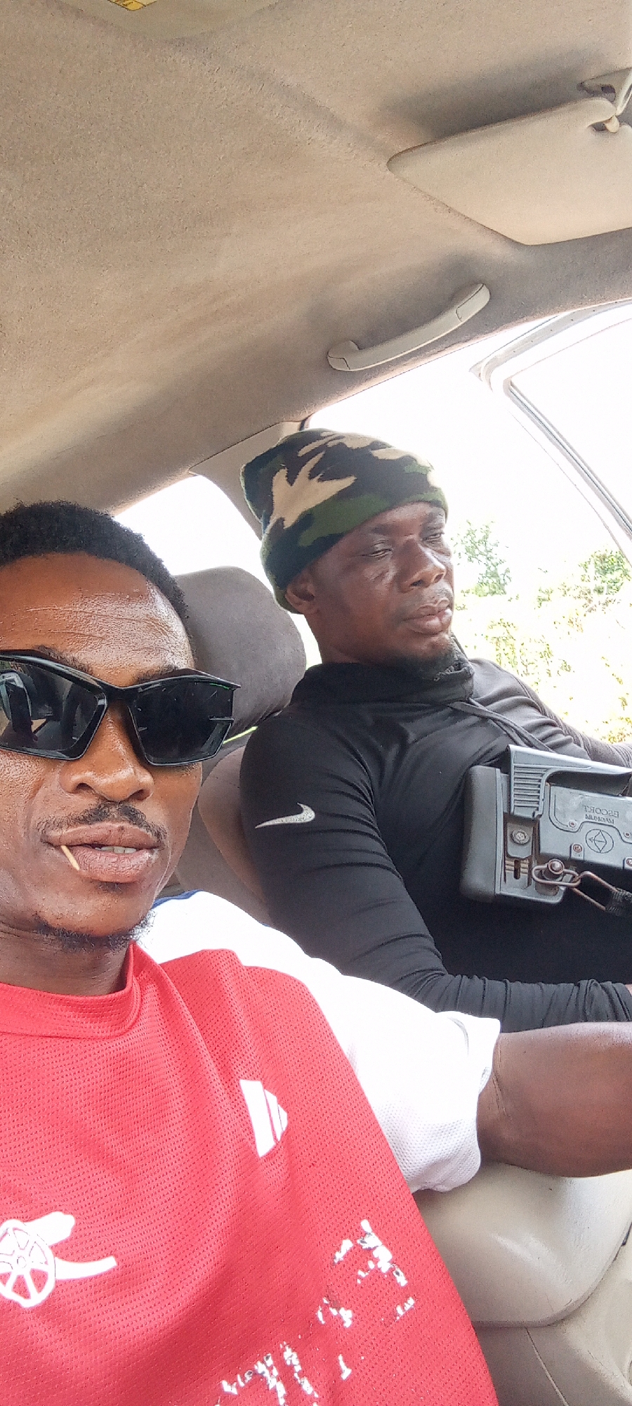 Two men are seated inside a car, captured in a selfie-style photograph, likely in Otunade, Nigeria. The man in the foreground wears large black sunglasses and a red Arsenal Football Club jersey featuring a white cannon logo. A toothpick is visible in his mouth.
Beside him, the second man wears a black long-sleeved shirt adorned with a white Nike swoosh logo on the sleeve, and a camouflage beanie. He appears to be resting, with his eyes partially closed. A dark object, resembling the stock and receiver of a firearm or specialized equipment, rests on his lap or chest. Clearly visible text on this object includes "ESCORT" and "MAGNUM," along with other small, illegible markings.
The car's interior, including the beige headliner, a sun visor, and a grab handle, is visible above them. Bright natural light streams through the car window, indicating a daytime setting, possibly sunny. Blurred greenery is visible outside the window, suggesting an outdoor, possibly natural or rural, environment. Both individuals maintain neutral expressions.