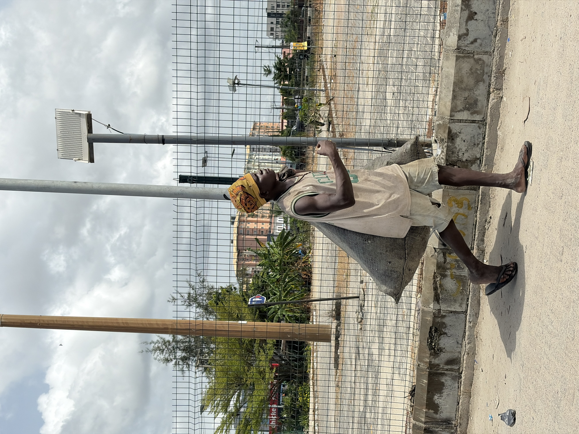 An African man sits on a gray sack alongside a wire fence, positioned between a concrete curb and a sidewalk in Igboefon, Nigeria. He wears a tan basketball jersey, khaki shorts, and brown sandals, with a patterned yellow headwrap. A light pole stands nearby, and buildings and foliage are visible in the background behind the fence.