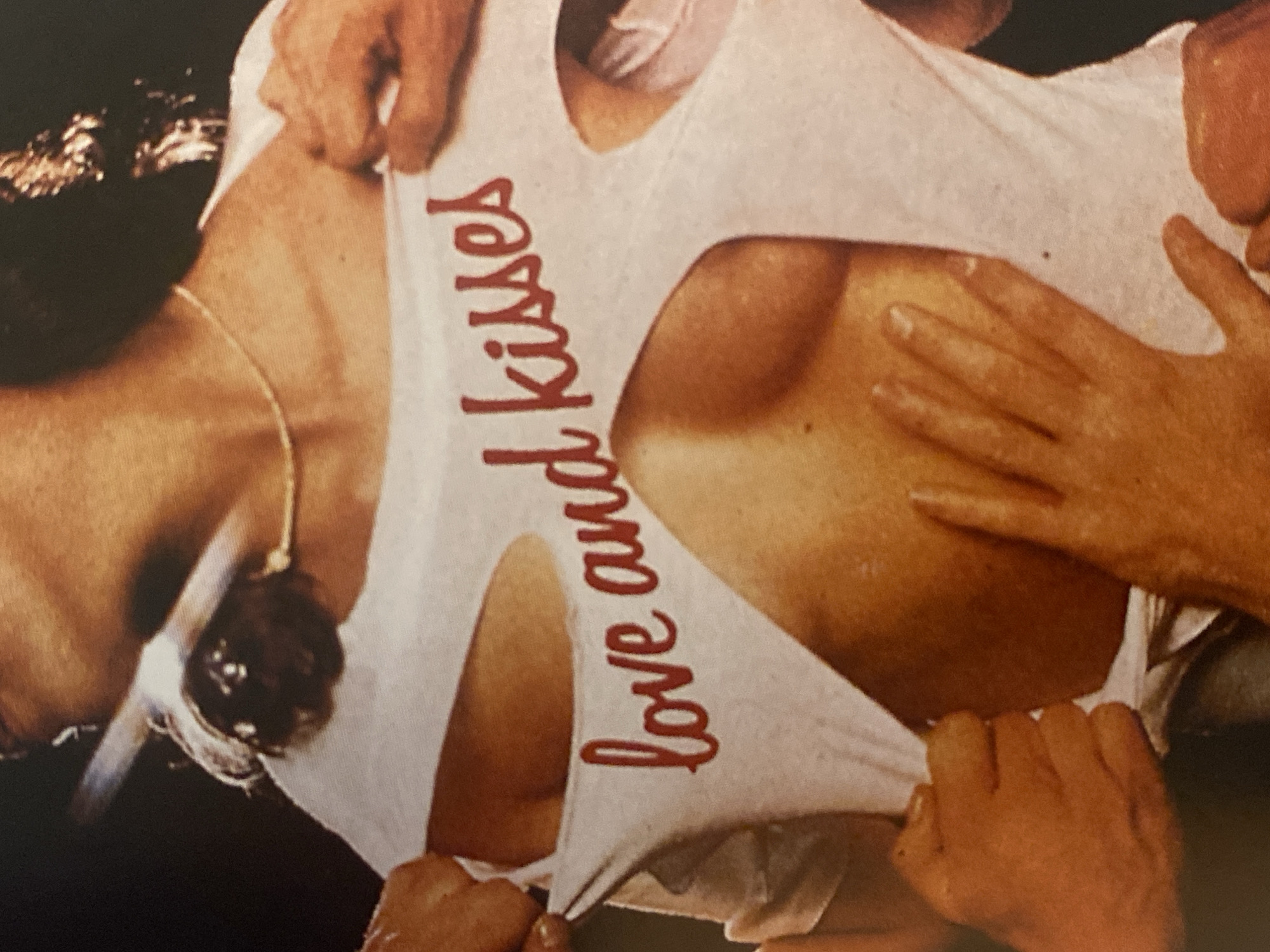 The image shows a person with a fair complexion and light brown hair wearing a white t-shirt with the words "love and kisses" printed vertically in red. The t-shirt is being pulled apart by multiple hands with fair skin. A gold chain necklace with a dark, round pendant is visible on the person's neck. The person's chest is exposed due to the torn t-shirt. One hand is resting on the person's upper chest. The background is blurry and out of focus. There is no indication of a location in the city of Шебунино, Russia.