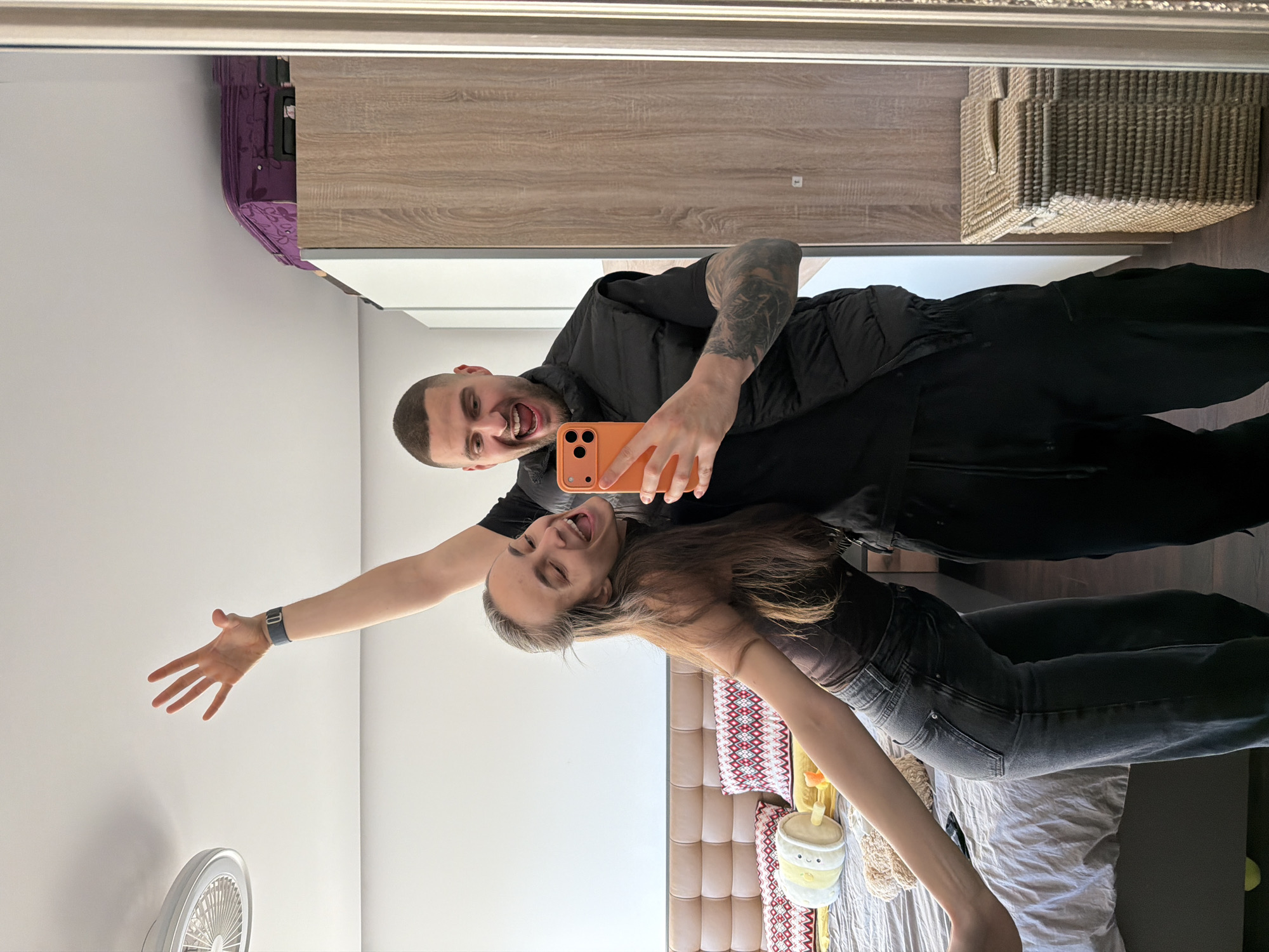 A man and woman are taking a selfie in front of a mirror. The man is in the foreground holding an orange phone and is wearing a black shirt and vest. He has a shaved head and tattoos on his arm. The woman is in the background with her tongue sticking out and her arm raised. She is wearing a brown shirt and black jeans. The background shows a bed with a decorative headboard and patterned pillows. The room also contains a wooden wardrobe, a woven basket, and a white wall with a fan. It's not possible to determine the exact location in Sofia, Bulgaria from the image alone.