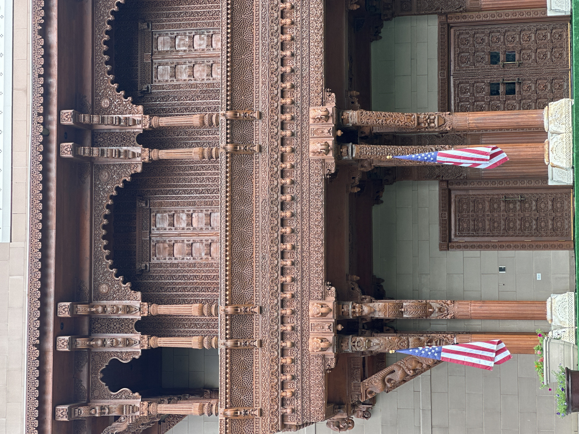 The image shows the exterior of a building featuring intricate wooden carvings, likely of Indian or South Asian architectural influence. The facade is characterized by a balcony with ornate wooden railings, pillars, and decorative friezes. Two American flags are displayed, one on each side of the central structure, hanging from poles. The flags are positioned in front of large, carved wooden doors. The building's walls appear to be made of light-colored stone or brick. Planters with flowers are visible at the base of the structure, suggesting an outdoor setting. The overall scene conveys a sense of grandeur and cultural fusion. The location is identified as Bartlett, United States of America.