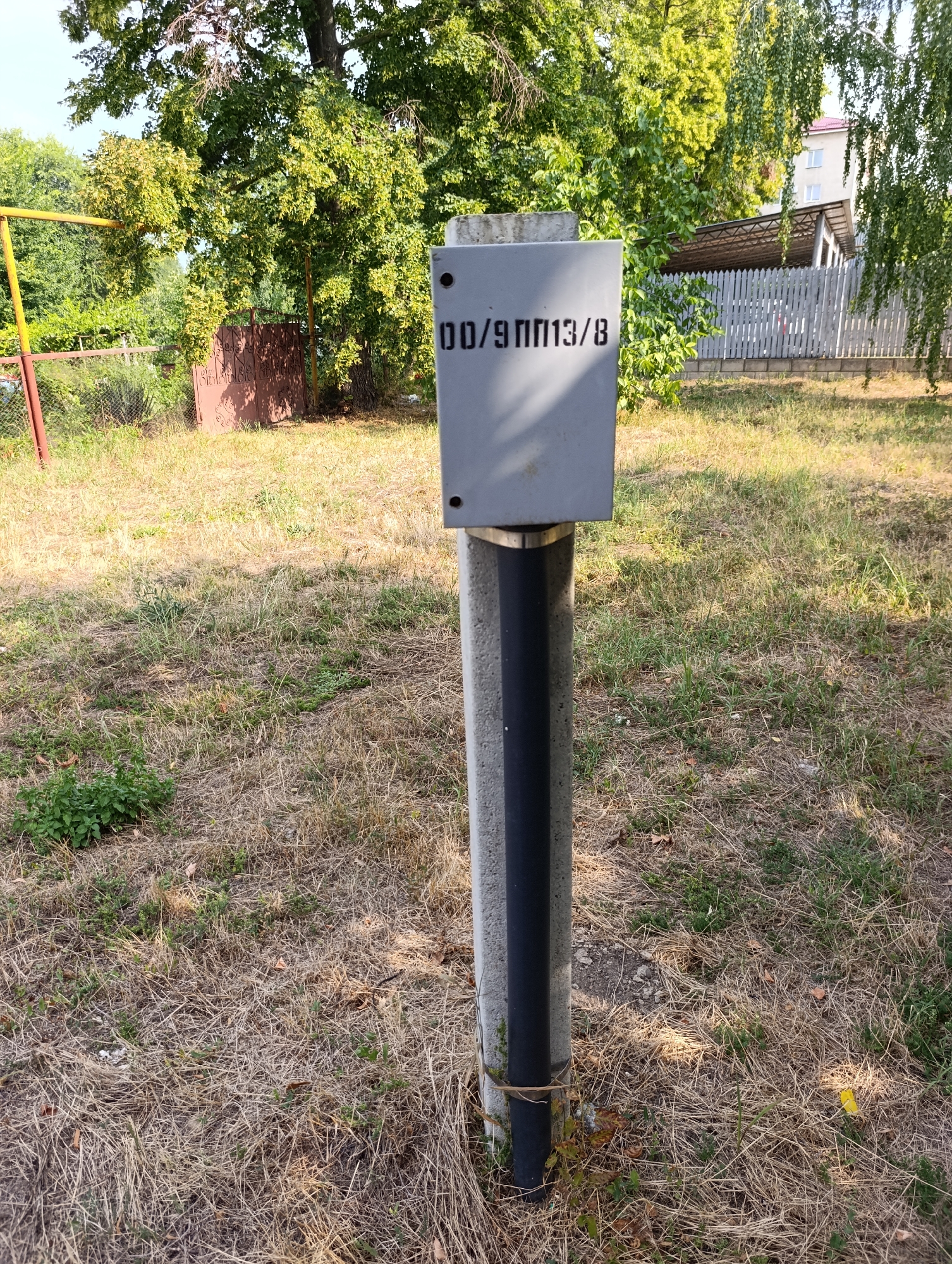 A gray electrical box with black lettering that reads "00/9ПП13/8" is mounted on a gray concrete pole with a black stripe. The box is positioned in an outdoor environment with dry, yellowed grass and some green vegetation. In the background, there are trees, a chain-link fence, a metal gate, and a building with a white fence. The scene appears to be in a park or a residential area. The location is the city of Dubăsari, town council, Moldova.