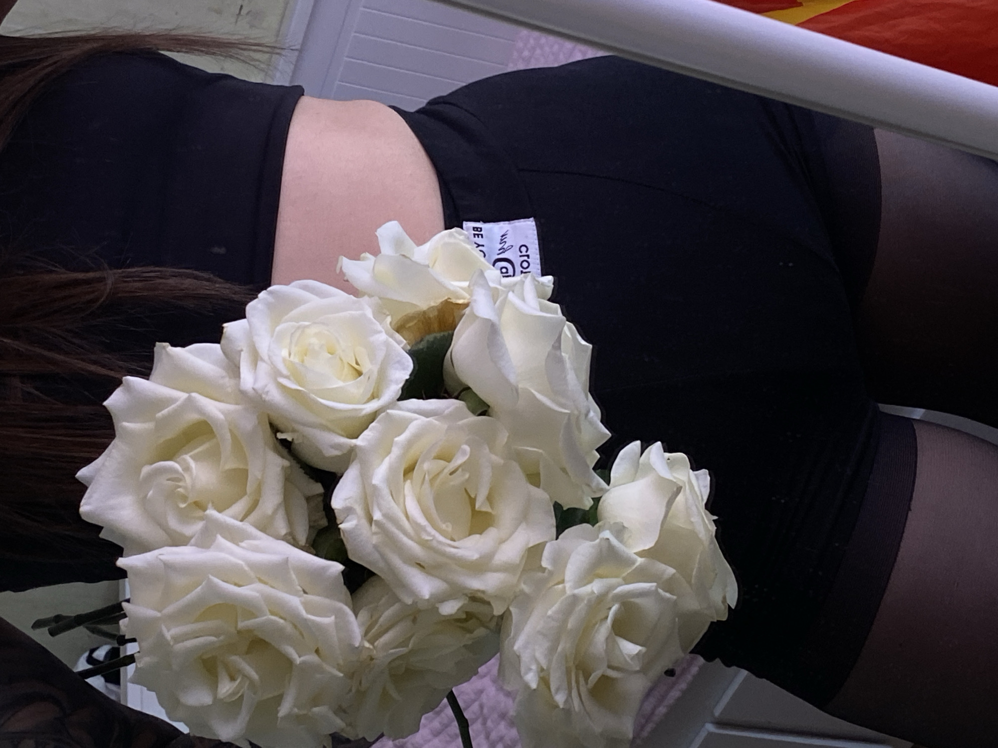 The image presents a close-up, high-angle view of a person lying down, partially obscured by a prominent bouquet of white roses. The individual wears a black crop top or bra and black shorts or underwear, complemented by dark, possibly sheer, stockings or tights. Long, dark hair cascades across the left side of the frame. A white tag on the black shorts is visible, showing fragmented text that includes "CROP" and "BE A".
A cluster of fresh, white roses with tightly packed petals dominates the foreground and central part of the image. The background is subtle, featuring a light-colored surface, possibly a bed frame or wall, and a hint of red and orange fabric in the upper right corner. The scene appears to be set indoors. The image seems to be a carefully composed photograph, possibly for aesthetic purposes. There are no direct cues about the time of day or weather. The implied location context is Votkinsk, Russia.