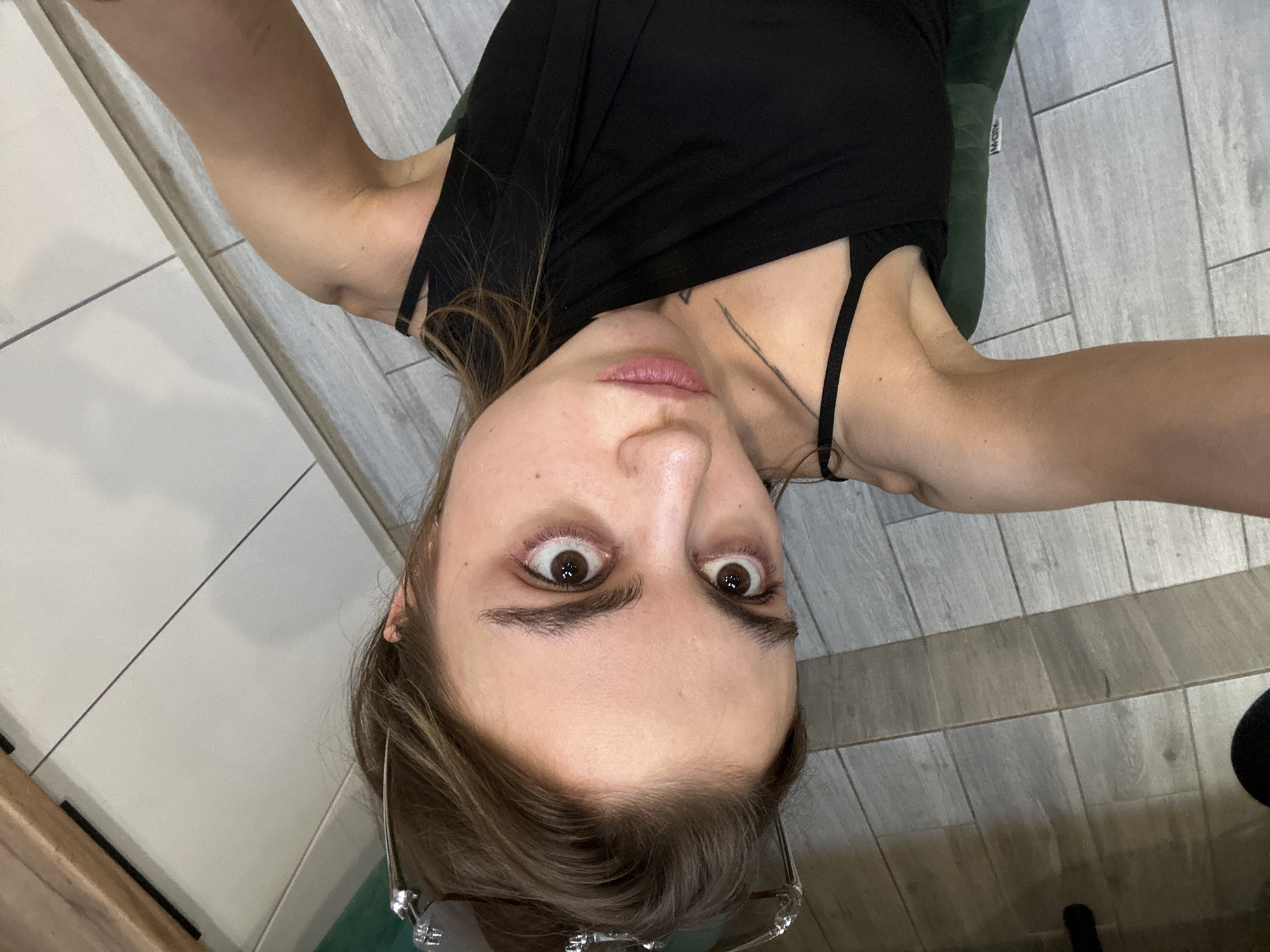 A young woman with brown hair and brown eyes is taking a selfie. She is wearing a black top with thin straps. She has light makeup on, with noticeable eyeliner and blush. There is a tattoo on her collarbone. She is lying down, and the camera is positioned above her. The background appears to be a bathroom with tiled floors and cabinets. The image is taken indoors. No specific location in Moscow, Russia can be identified.