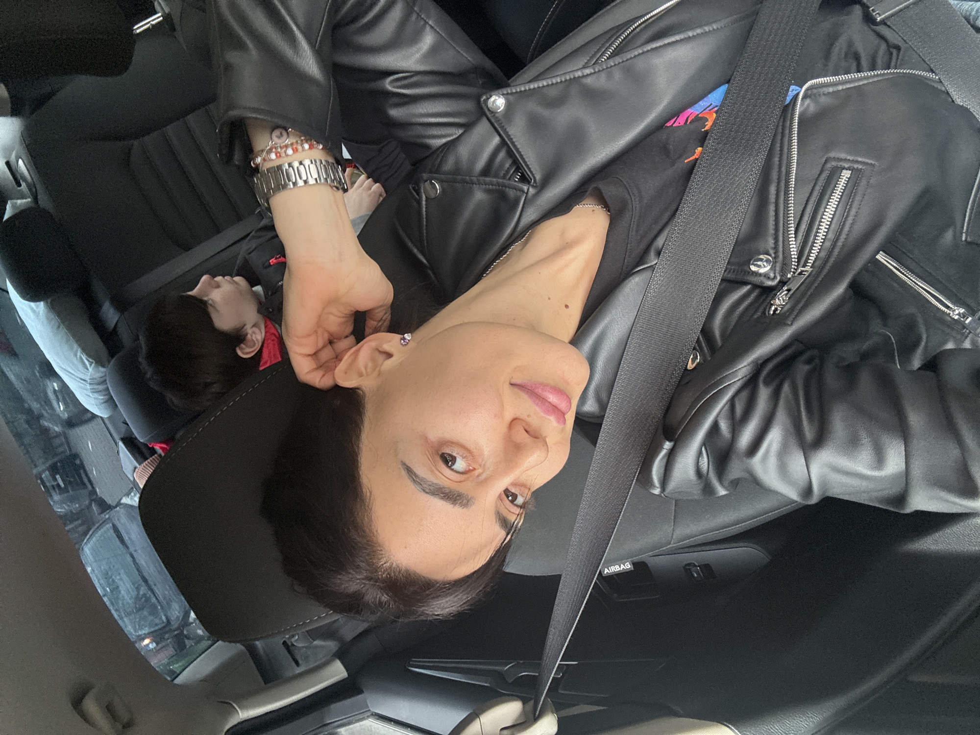 A woman wearing a black leather jacket and multiple bracelets is sitting in the driver's seat of a car, wearing a seatbelt. A child lies in the back seat behind her. The car appears to be in Tbilisi, Georgia, based on the street scene visible through the car windows.