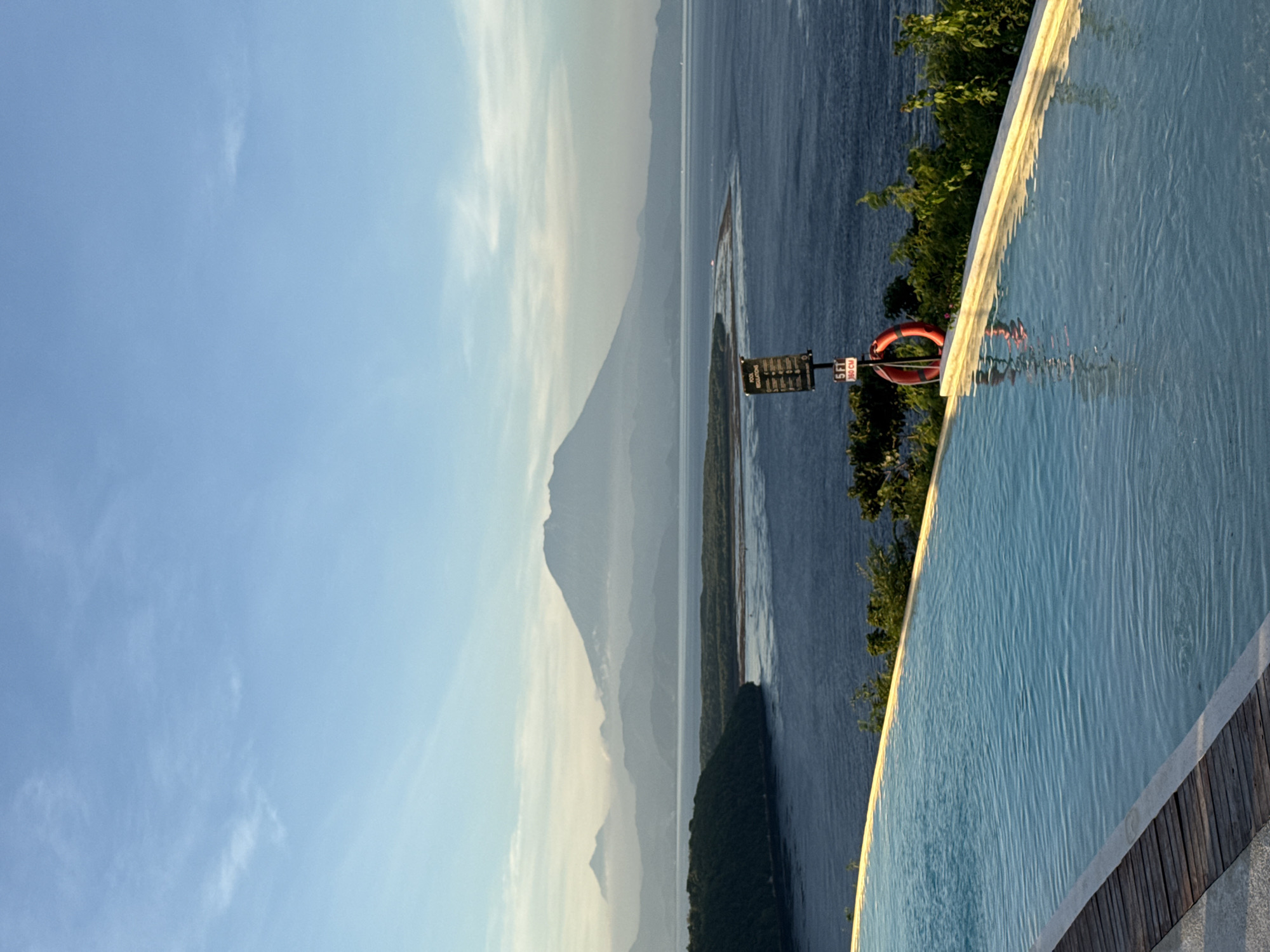 The photograph depicts a scenic view from a high vantage point on Nusa Penida, Indonesia. In the foreground, the edge of an infinity pool is visible, featuring light blue water with gentle ripples. Adjacent to the pool are wooden planks. Beyond the pool, lush green vegetation grows on the edge of the cliff. Further in the background, a large body of water extends towards a mountainous island. The sky is clear blue with some light clouds. Near the edge of the pool, a small sign with regulatory information and a red life preserver are mounted on a post. The overall setting suggests a luxurious resort or private residence overlooking the ocean.