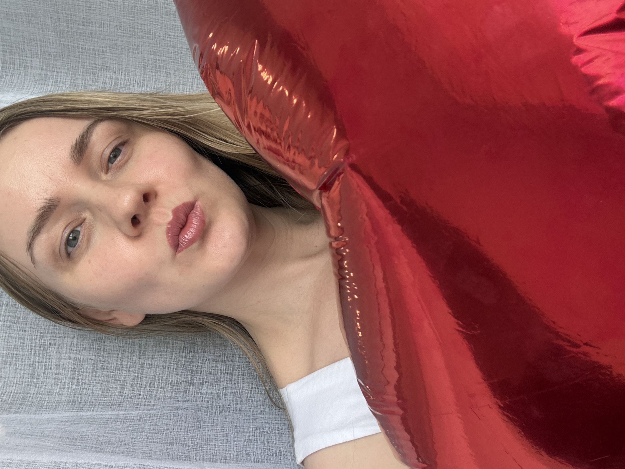A woman with blonde hair is positioned next to a large, red, heart-shaped foil balloon. She is wearing a white top. The background appears to be a gray, textured surface, possibly fabric. The location is not indicated by any identifiable features in the image.