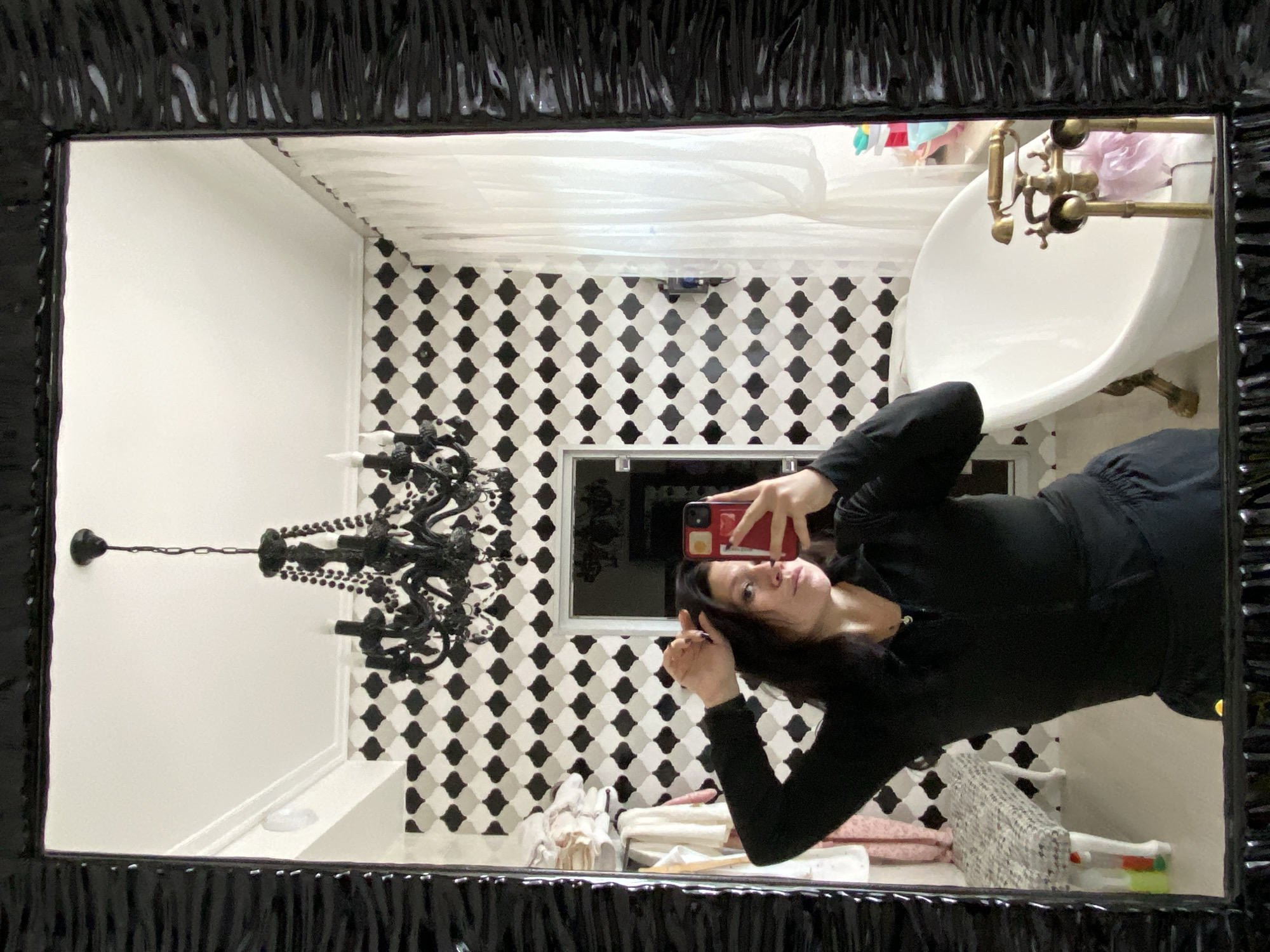 A woman is reflected in a mirror in what appears to be a bathroom. She is wearing a black long-sleeved top and black pants. She is holding up a red smartphone to take the picture. The bathroom has white walls and a black and white patterned tile backsplash. There is a clawfoot bathtub with brass fixtures. A black chandelier hangs from the ceiling. The reflection is oriented upside down in the mirror. The overall aesthetic suggests a somewhat gothic or dramatic decor. The location is provided as "Прикамский, Russia," which is a region in Russia. There is no visible text in the image.