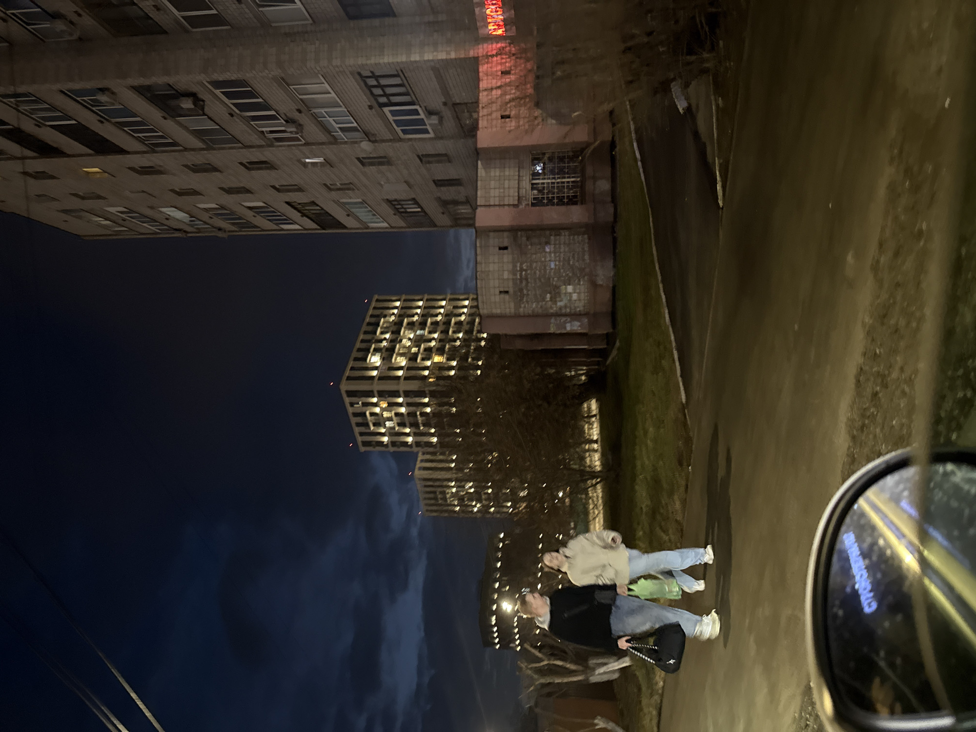 The image depicts an evening scene in Izhevsk, Russia. Two people, a female in a black jacket and the other in a tan jacket, are walking on a sidewalk. Tall buildings, including residential blocks with lit windows, are visible in the background, a street with a curb, and grass in the foreground. A car's side mirror is visible in the lower-right corner of the image, suggesting the photo was taken from inside a vehicle.