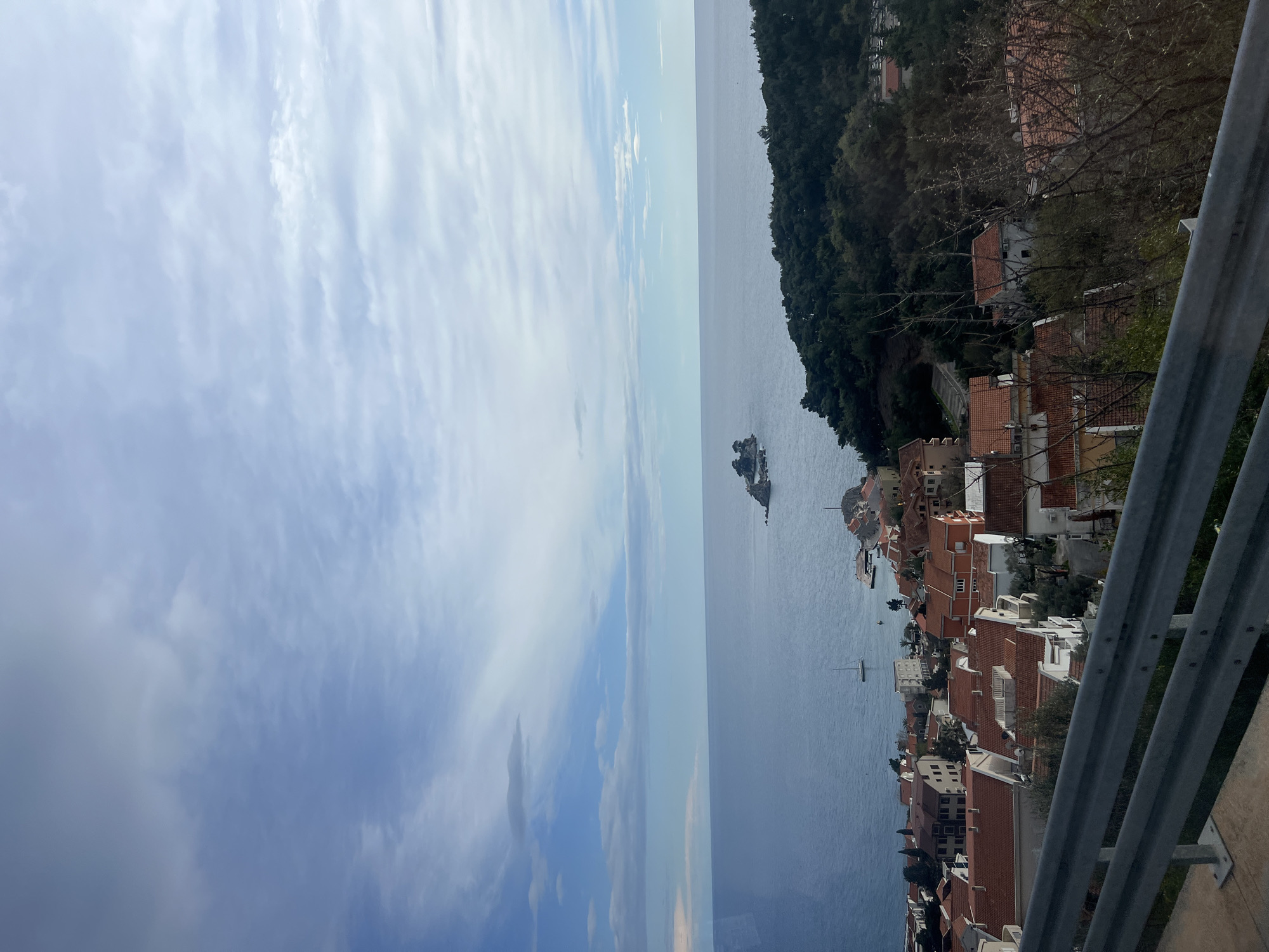 The image captures a coastal scene in Petrovac na Moru, Montenegro. The view is angled, featuring a landscape with a mix of natural and urban elements.
The Adriatic Sea stretches out horizontally, meeting a horizon with a partly cloudy sky. A small island sits just off the coastline.
On the right, a dense cluster of buildings with red-tiled roofs is visible, nestled amongst trees and vegetation on a steep hillside. The architectural style appears Mediterranean.
A metal guardrail runs along the bottom of the frame, suggesting the view is taken from an elevated position, possibly a road or overlook.
The image has a somewhat grainy quality. There are no people visible in the frame. The overall impression is of a scenic overlook of Petrovac's coastline.