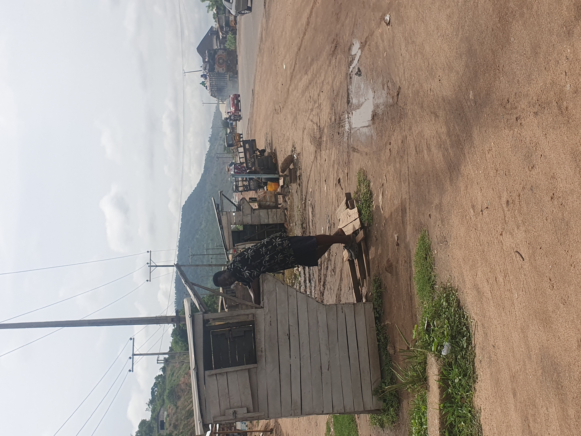 The image shows a man leaning on a wooden structure, likely a small shop or booth, on a dirt road in a rural or developing area possibly located near or within Lokoja, Nigeria. The man is wearing a dark-colored patterned shirt and dark shorts. The wooden structure appears weathered and is constructed from planks. Behind the structure, electrical power lines stretch across the frame. Further back, there's a road with some vehicles, and a hill or small mountain is visible in the distance under a cloudy sky. The ground is mostly dirt with patches of grass and weeds.