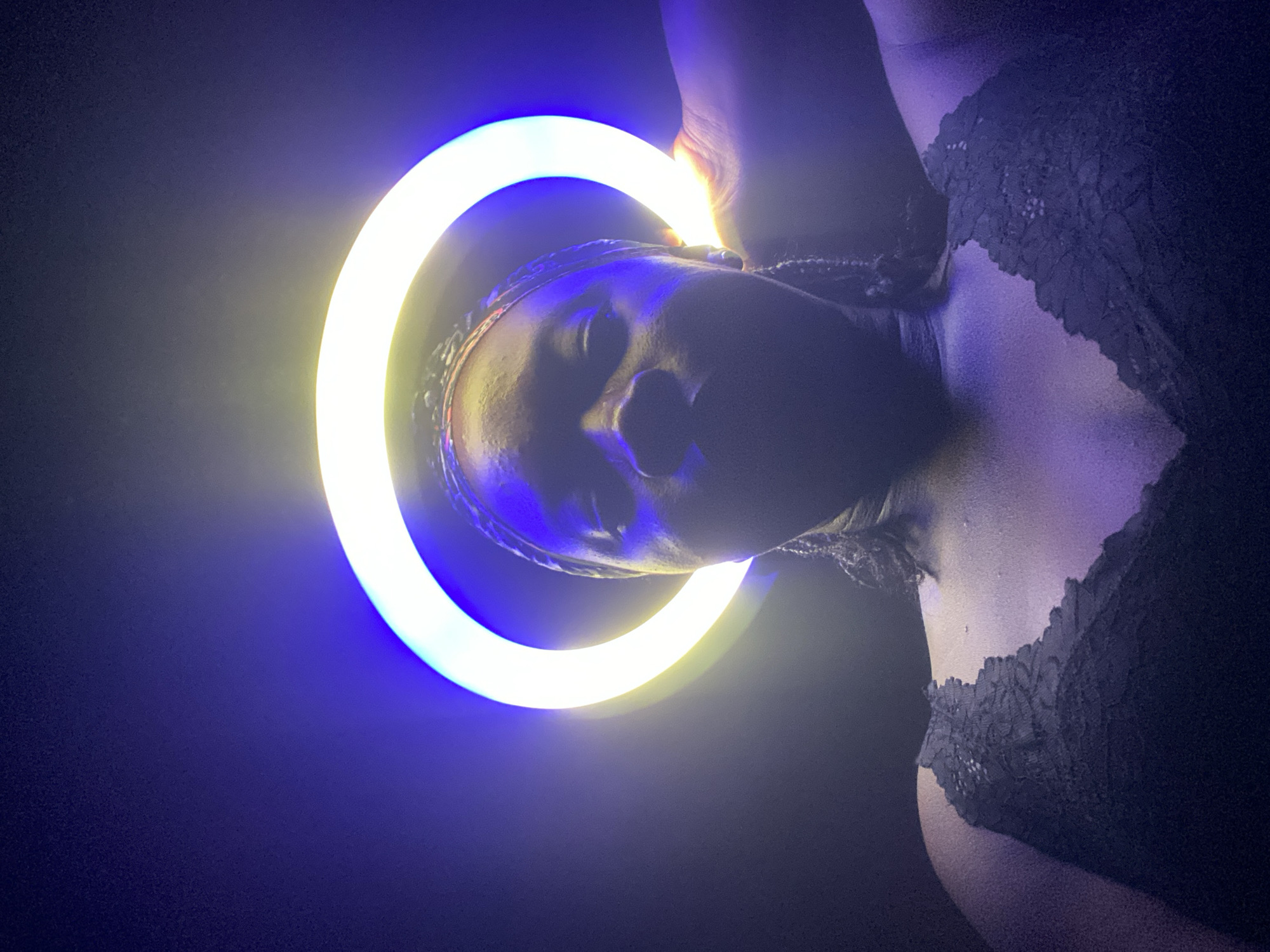 A person with their face illuminated by a blue and yellow ring light, wears a dark, lacy garment. The scene is dark, with the face tilted towards the camera. The person is holding the light. The location is unknown, but mentioned to be Maigizo, Nigeria.