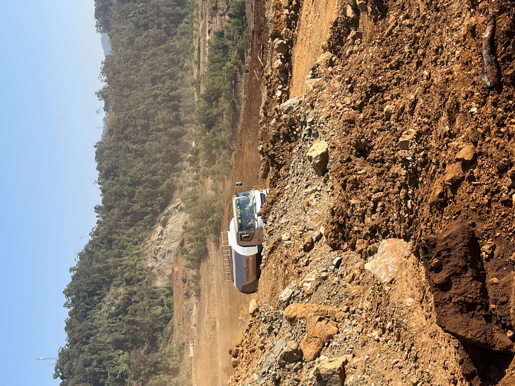An over-the-shoulder shot captures a white fuel truck driving through a barren, construction site in a mountainous, forested region of Indonesia. The truck, bearing the text "PT SUMBERBAHANA MINERAL" and "20 FUEL," is positioned on a dirt path, kicking up dust. In the background, lush green trees cover the hillsides, with some areas of deforestation visible, indicating ongoing development or resource extraction. The foreground is dominated by piles of reddish-brown soil and rocks. The sky is a clear, bright blue.