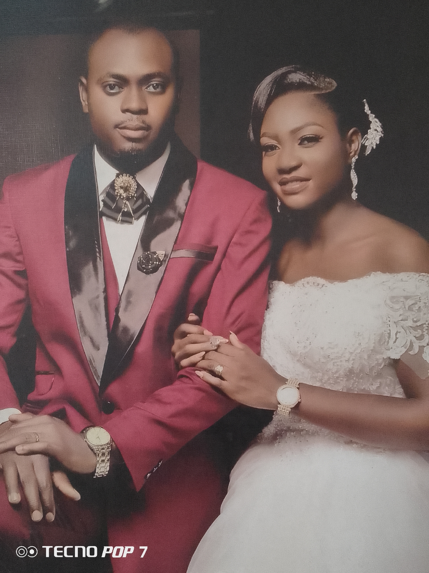 A wedding portrait shows a couple against a dark backdrop. The man wears a maroon suit with a black satin lapel, a white shirt, and a decorative bow tie. He also wears a wristwatch and a wedding ring. The woman is in a white, off-the-shoulder wedding gown, with her hair styled up and adorned with a decorative piece and earrings. She also wears a wristwatch and multiple rings. Their hands are clasped together. The photo appears to have been taken with a TECNO POP 7 phone, indicated by the watermark at the bottom. Location is unknown.