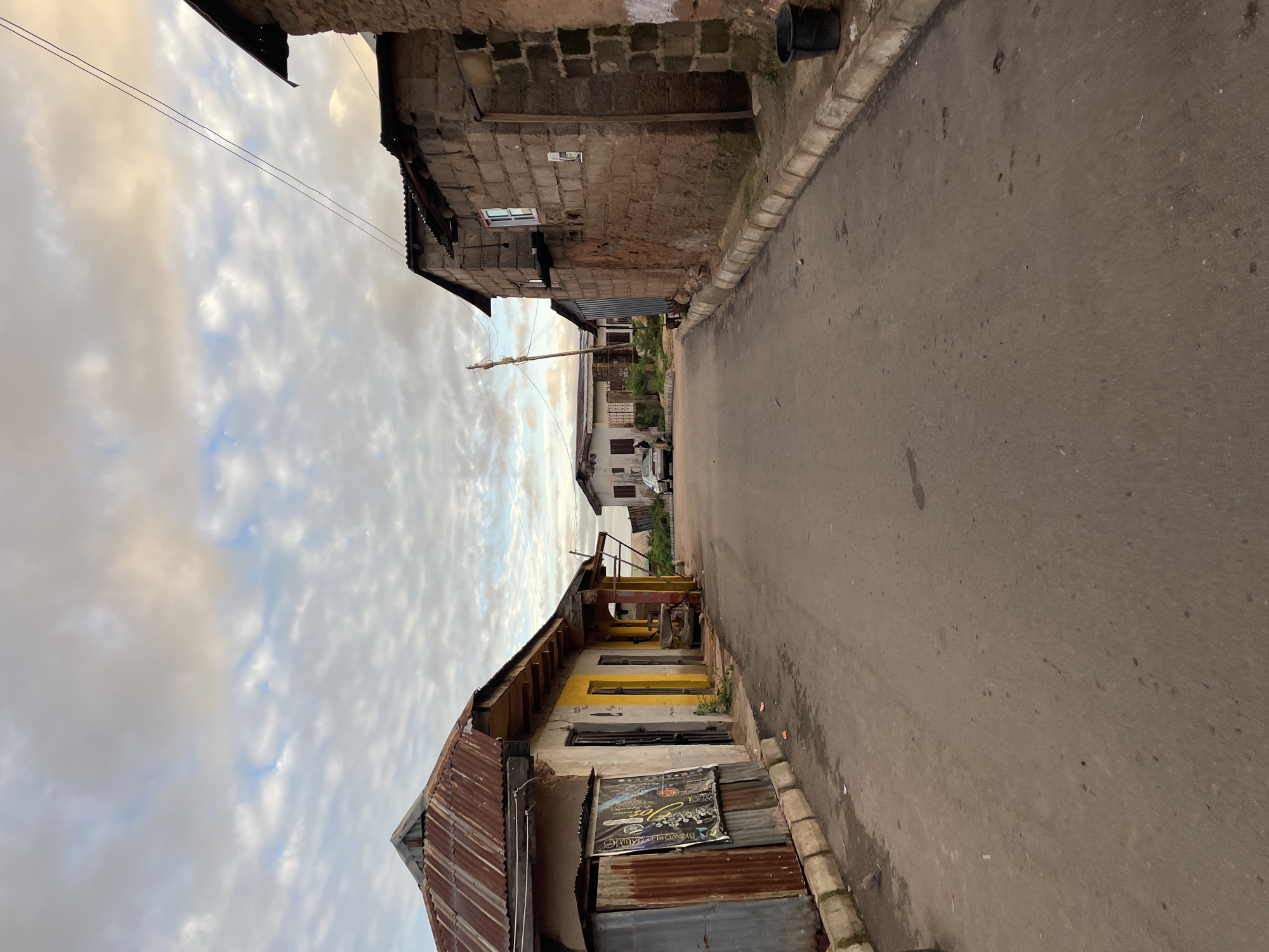 A street scene in Jos, Nigeria, shows a paved road with buildings lining both sides. The architecture is a mix of corrugated metal roofs and concrete block structures, suggesting a developing urban environment. The sky is partly cloudy, with a soft, diffused light indicating either early morning or late afternoon. On the left, a building with a rusted corrugated metal roof has a partially visible poster attached to its facade. Yellow and gray paint is visible on this building. On the right, a more robust building constructed from rough-hewn concrete blocks stands. A black bucket is placed near the curb on the right side. In the distance, a silver car is parked on the side of the road, and a person can be seen near it. The overall impression is one of everyday life in a residential or commercial area of Jos.