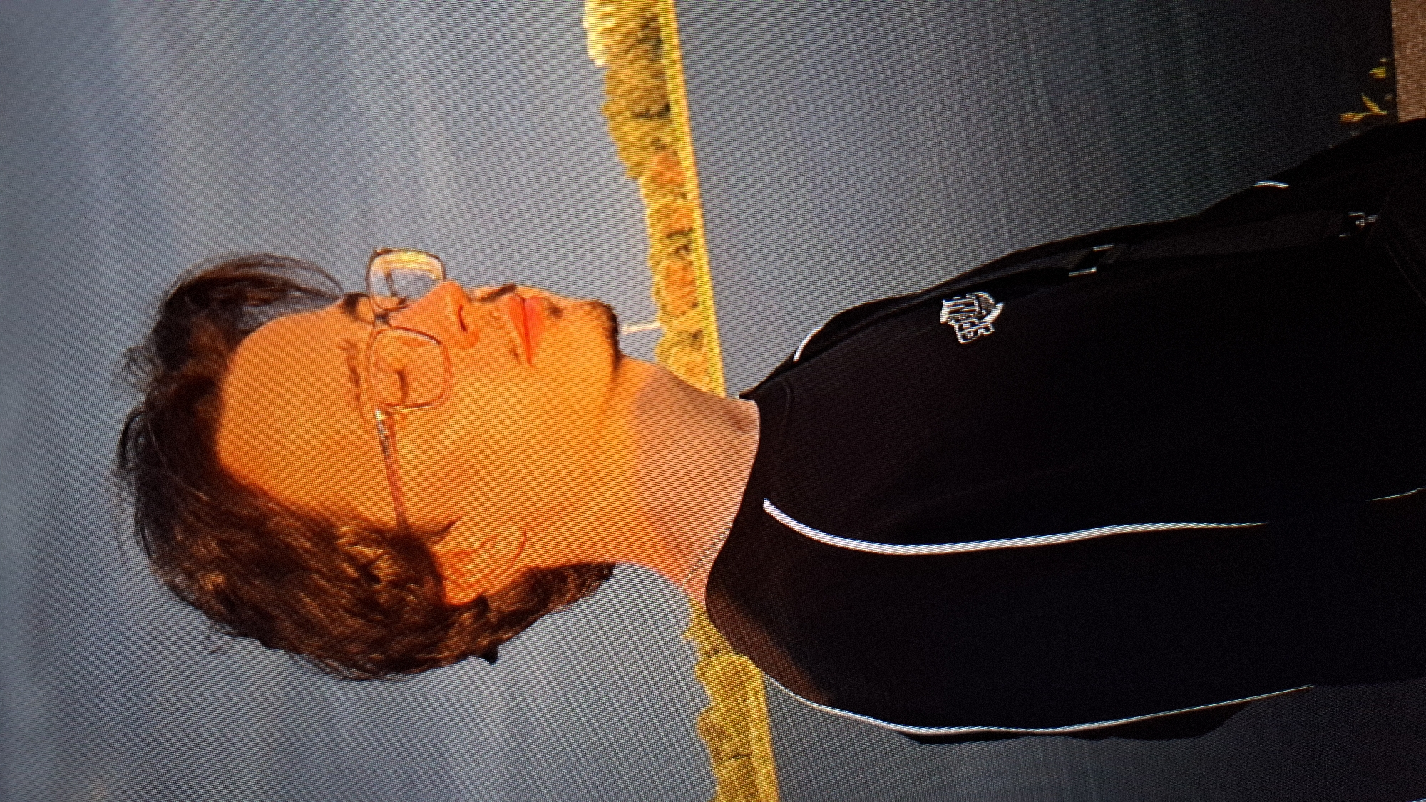 The media file shows a close-up, vertically oriented shot of a young man from the chest up. He has dark, wavy hair and is wearing glasses with thin wire frames and clear lenses. His head is tilted back, and his eyes are closed, giving him a relaxed or contemplative expression. The lighting suggests it is either sunrise or sunset, casting a warm, orange glow across the left side of his face, highlighting his nose, lips, and cheek. He appears to have a light beard or stubble. He is wearing a black crewneck sweatshirt with a white trim along the sides and a small, metallic logo on the left breast, which appears to be an emblem of some kind. The background is out of focus, showing a muted blue sky and a blurred yellow and brown textured line that could be a distant object or structure. The scene seems to be outdoors, possibly in a park or open area in Balakovo, Russia, given the location context, with the time of day being either early morning or late evening due to the strong directional light.