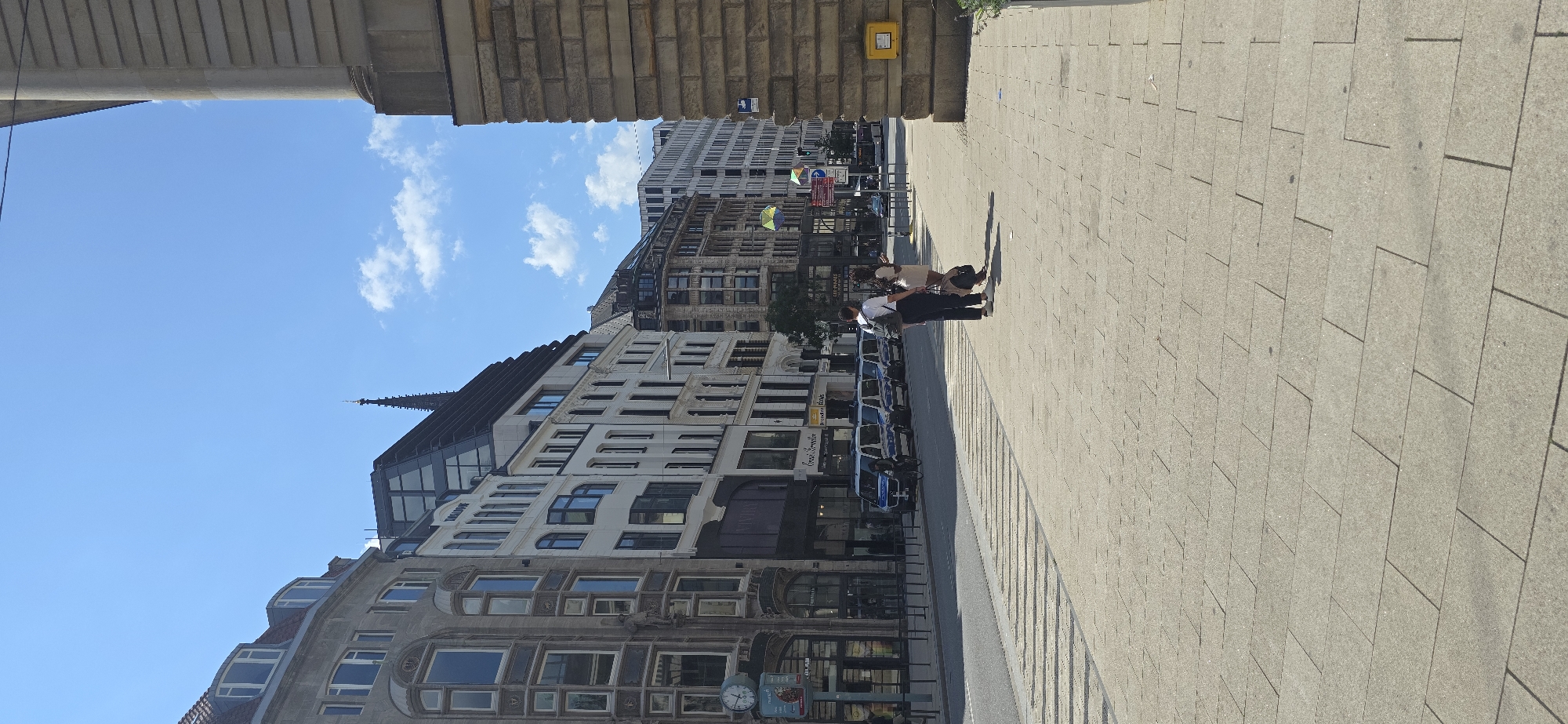 The image shows a street scene in Hamburg, Germany. A street is paved with light-colored rectangular stones. On the left side of the street, several multi-story buildings line the sidewalk. These buildings vary in architectural style, with some featuring older ornate facades and others with more modern glass and dark roofing. A clock is visible on the facade of one of the buildings.
On the right side of the street, a large, light-colored stone building with horizontal banding is prominently featured, partially obscuring the view of other buildings behind it.
In the street, a line of white vans, possibly police vehicles due to their markings and presence, are parked. A person on a bicycle is visible in front of the vans. Two individuals, a man and a woman, are standing on the sidewalk, facing away from the camera and towards the street. The man is wearing a backpack and the woman is carrying a bag. They appear to be conversing.
The sky is blue with scattered white clouds. The overall scene suggests a clear, sunny day in an urban environment.