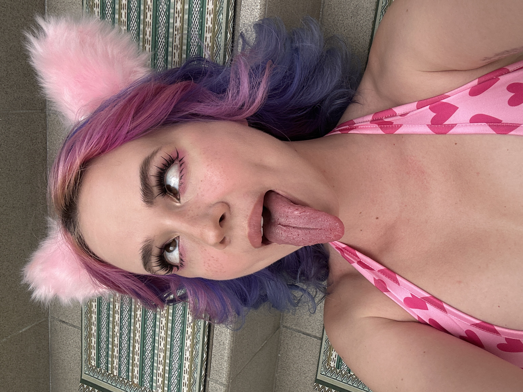 A woman with pink and blue hair and pink furry cat ears is lying on her side, sticking her tongue out. She is wearing a pink top with red hearts. She is lying on a patterned mat on a concrete surface. The exact location in the City of Zagreb, Croatia is indeterminable from the image.
