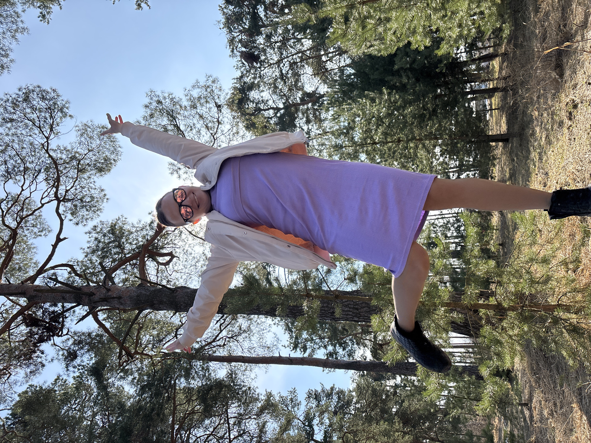 A young female stands in a pine forest in Раздолье, Russia, captured from a low-angle perspective. She is positioned centrally, smiling at the camera while wearing rose-tinted sunglasses, a light purple ribbed midi-dress, and a light cream jacket with an orange lining. Her arms are raised and outstretched, palms open, and her left leg is lifted and bent at the knee, with the foot pointing left, while her right foot supports her on the ground. The background consists of numerous tall pine trees with green foliage, silhouetted against a clear blue sky. Bright sunlight emanates from behind and above the subject, causing lens flares. The forest floor is covered with dry leaves, pine needles, and sparse dry grass, with several smaller pine saplings visible.
