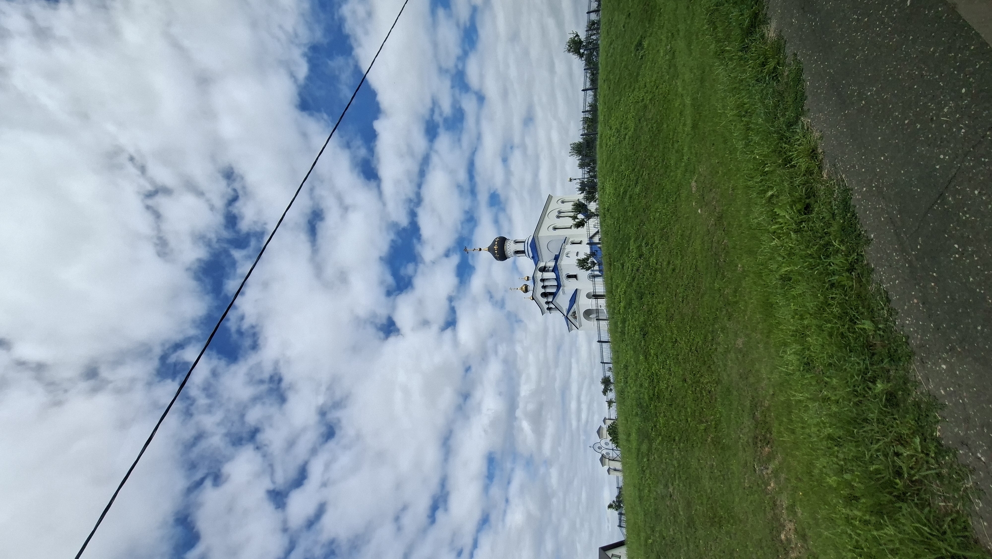 A white church with blue roofs and golden domes sits atop a grassy hill under a cloudy blue sky. A black wire runs diagonally across the sky. In the foreground, a paved area is visible, bordering the grassy slope. The church appears to be an Orthodox Christian church, judging by its architectural style. The overall scene suggests a rural or suburban setting in Myadzyel, Belarus. There are no people visible in the image.