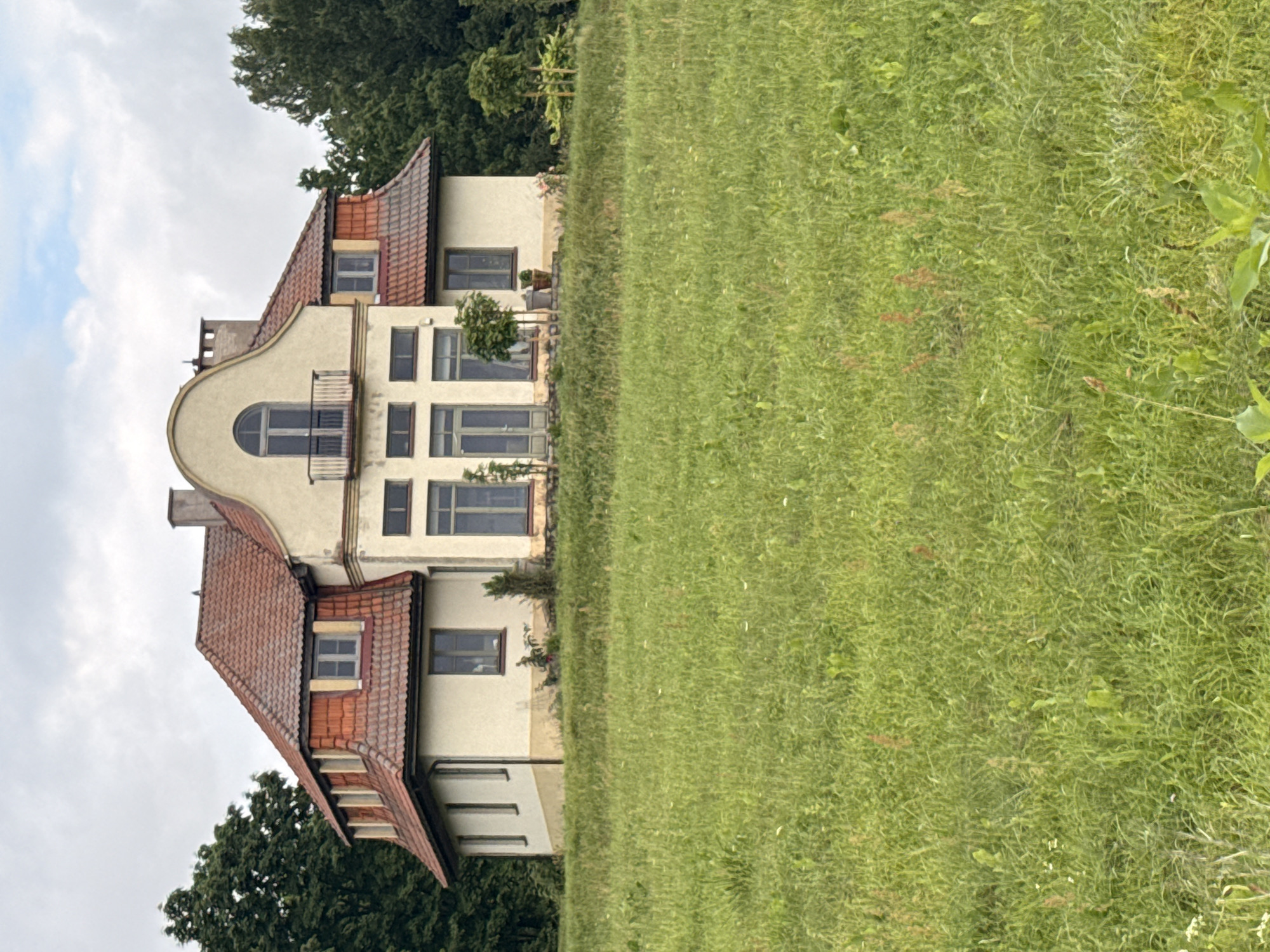 This is a full, eye-level, outdoor, vertical shot of a large, tan, two-story house with a red tile roof, set against a backdrop of lush green trees and a pale, overcast sky. The house is situated on a gentle slope, with the foreground dominated by a wide expanse of bright green grass speckled with wildflowers and some dry weeds. The house itself has a prominent mansard roof with several dormer windows. The front facade features a rounded arch window above a small balcony on the second story, flanked by two smaller windows. The first floor has a central section with tall windows and doors, and two smaller wings extending to the sides, each with rectangular windows. A chimney is visible on the right side of the roof. Several small trees and potted plants are placed around the base of the house. The area around the house appears to be a rural or semi-rural setting, possibly a park or estate, located in Aukštieji Rusokai, Lithuania. There are no people or animals visible in the image.