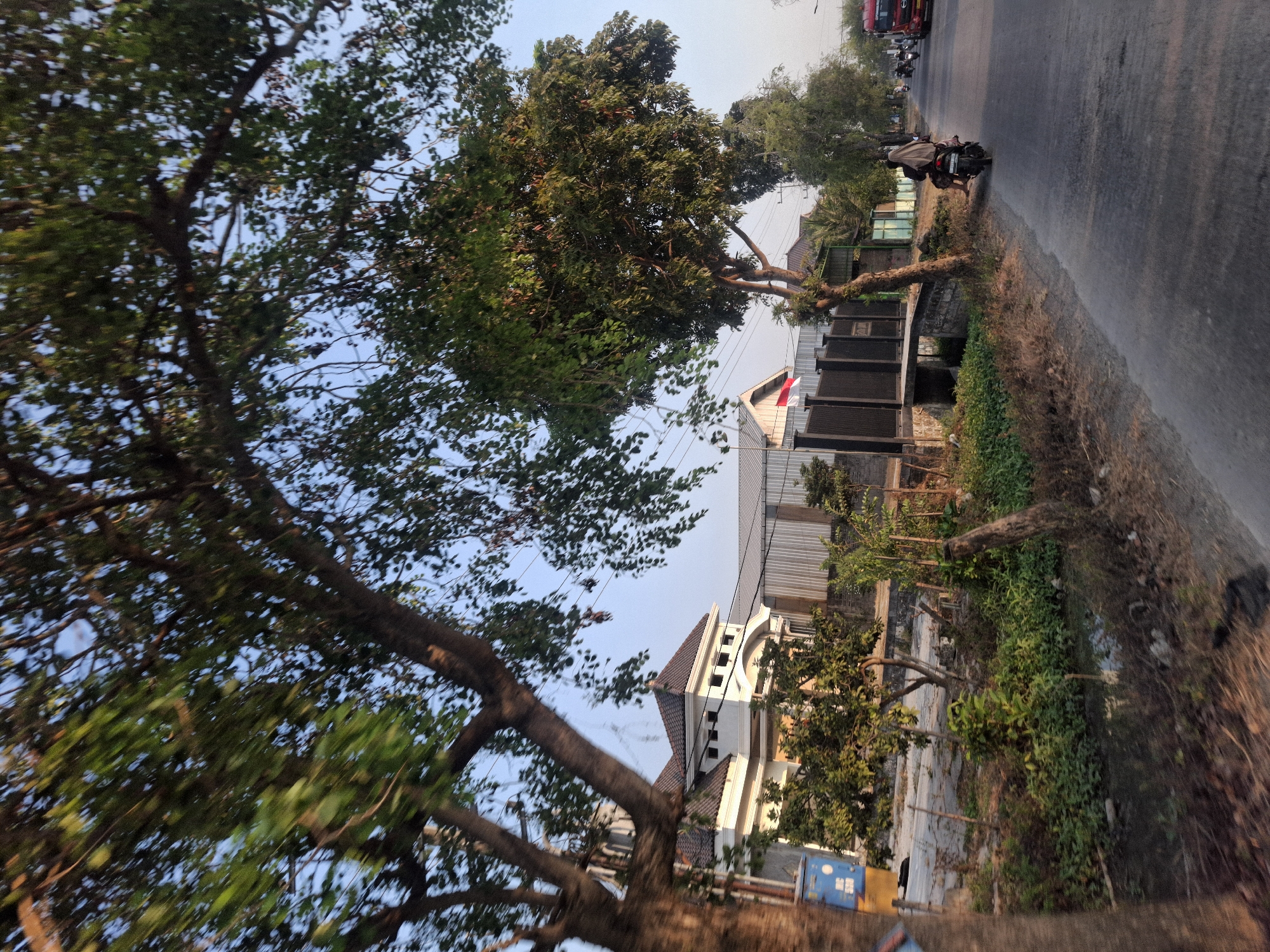 The image depicts a street scene in Banjarmadu, Indonesia, on a clear day. The foreground is dominated by the blurred motion of a vehicle, with a tree's trunk and branches partially obscuring the view on the left. A paved road stretches into the distance on the right.

Along the side of the road, there is lush vegetation, including small trees and bushes. In the middle ground, a modern white house with a dark-tiled roof is visible, featuring multiple windows and an arched entrance. Adjacent to the house, there's a fence and a corrugated metal building. A red and white Indonesian flag is mounted on a pole near the fence.

Further down the road, a motorcycle carries two people wearing traditional attire. Several other motorcycles are also seen on the road. A red truck is visible in the far distance on the right. The sky is a clear, pale blue. The overall impression is that of a suburban or semi-rural area.