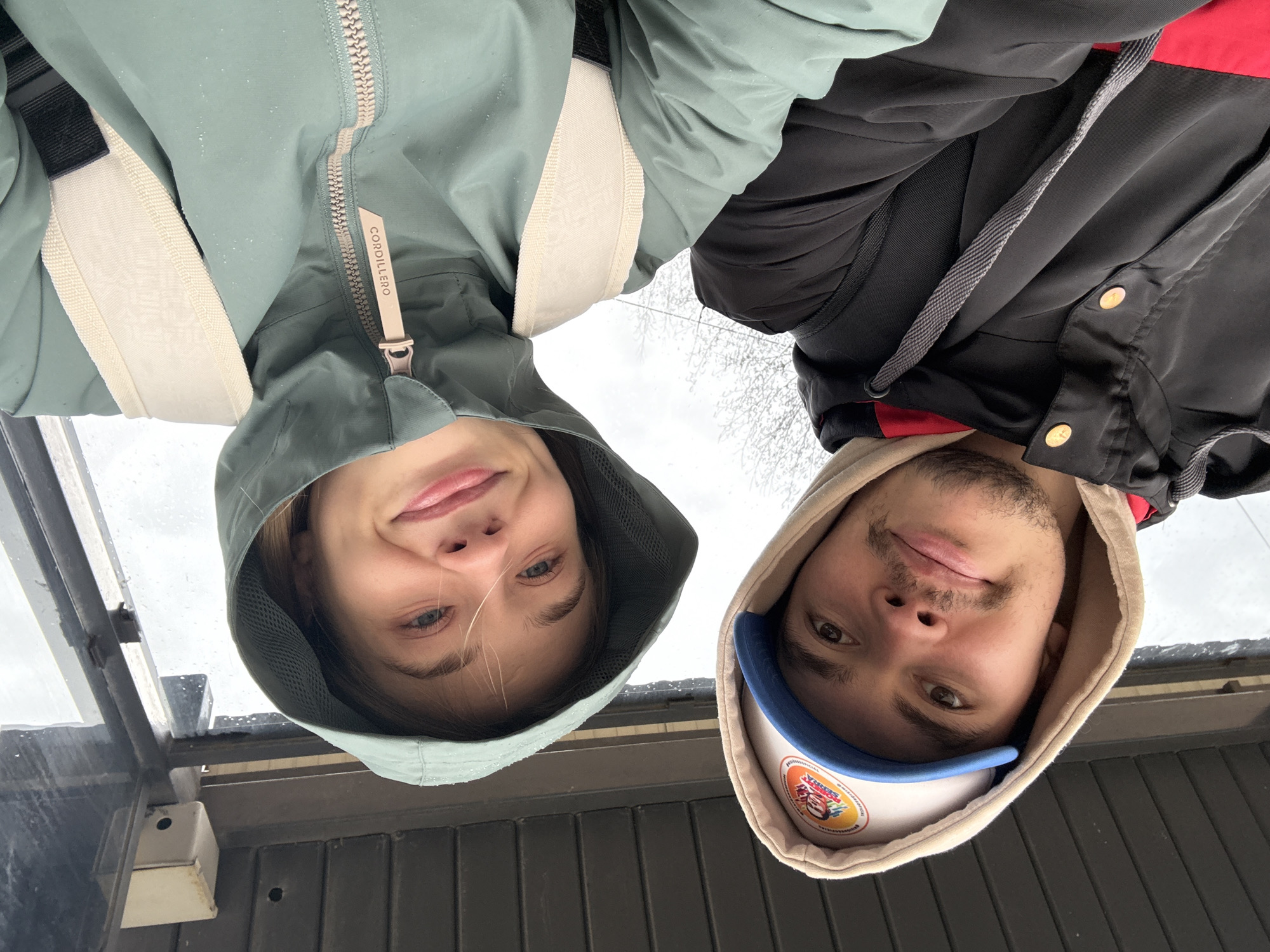 This media file is a selfie, rotated 90 degrees clockwise, featuring two young individuals looking upwards into the camera from a low angle. The setting appears to be outdoors, likely under a shelter or awning, as indicated by structural elements and a translucent panel with water droplets visible in the foreground (when correctly oriented). The sky in the background is overcast and light grey, with blurred bare tree branches suggesting a natural backdrop.
On the left, a person with light-colored hair is wearing a light teal/green hooded jacket with the hood up. The zipper pull on their jacket is clearly visible, displaying the brand name "CORDILLERO". They have a subtle, content smile. On the right, a young man with facial stubble is wearing a light beige hooded sweatshirt, with a black jacket featuring red accents visible over it. He also wears a white baseball cap with a blue brim underneath his hood and has a slight smile.
Both subjects convey a sense of calm or positive emotion through their gentle smiles. The presence of water droplets on surfaces, including potentially on the left person's jacket, suggests recent rain, mist, or melting snow, indicating cool or damp weather. The foreground also includes a dark, possibly metal or concrete, structure with vertical lines and some attached white box-like objects. The scene captures a casual moment between the two individuals, likely posing for a shared photograph in an outdoor, possibly urban or suburban, environment during an overcast day in Chaikovsky, Russia.