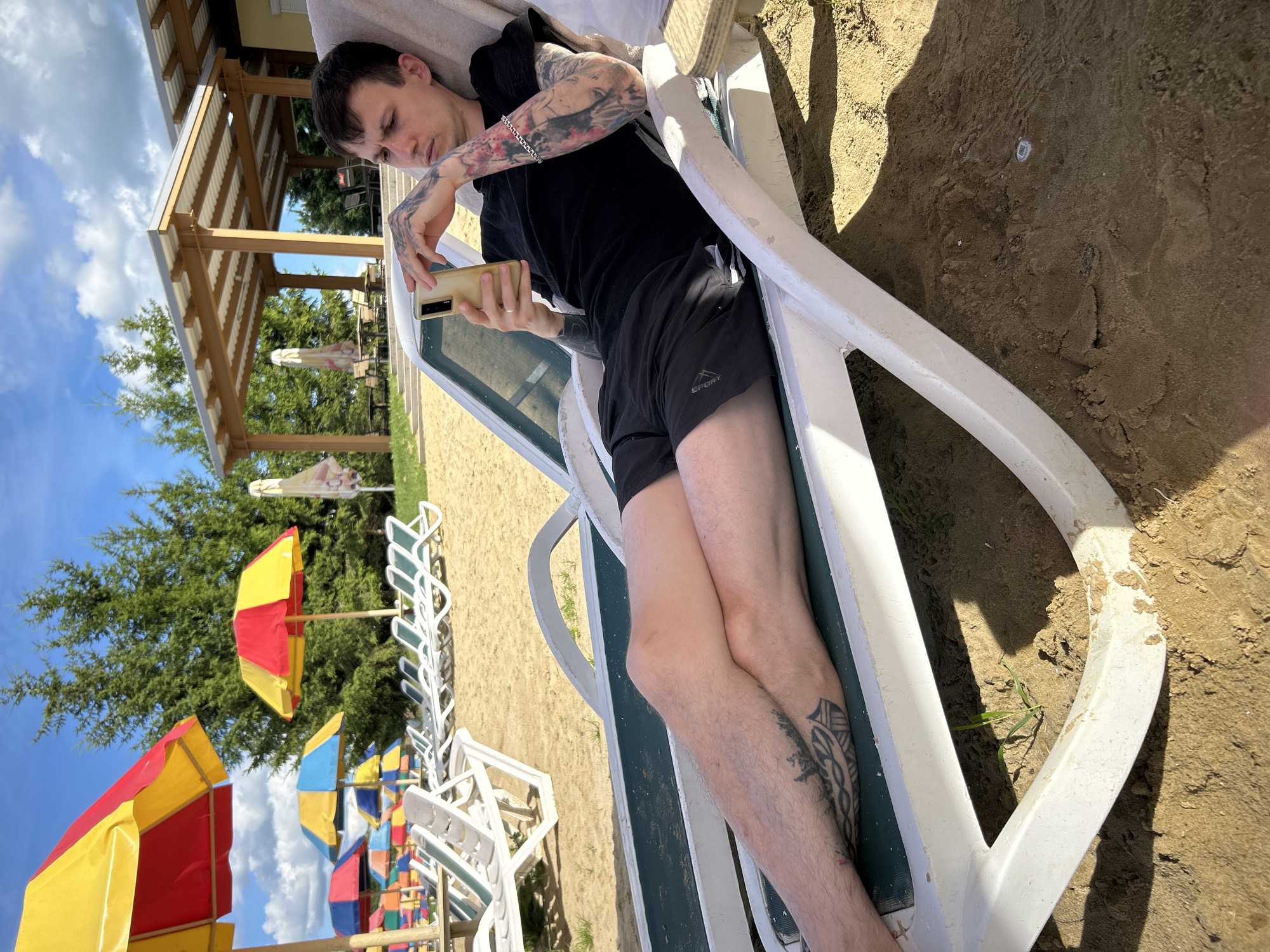 A young man with multiple tattoos on his arms and legs is relaxing on a white lounge chair on a sandy beach. He is wearing a black t-shirt and black shorts and is holding a smartphone in his hands, looking down at it. The background shows several beach umbrellas in red, yellow, and blue, and a wooden structure with a covered roof. Trees are visible behind the structure. The scene appears to be a recreational area, possibly a beach resort or park in the city of Yubileyny, Russia. The lighting suggests it is a sunny day with some clouds in the sky.