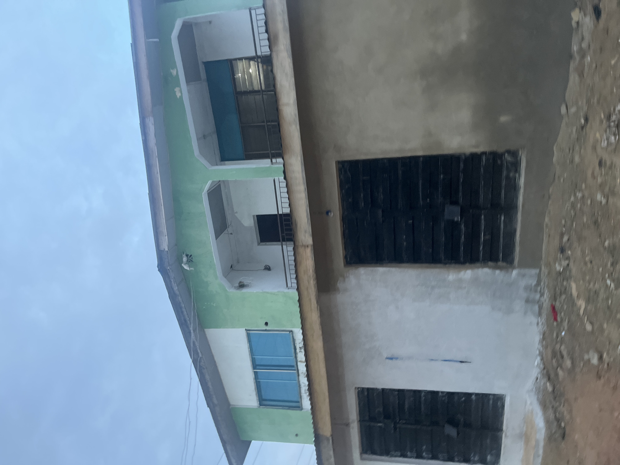 The image depicts a two-story building under construction or renovation in Ikorodu, Nigeria. The upper story is painted green and white, with blue tinted windows on the left side and a balcony with a metal railing on the right, featuring dark-tinted windows. The lower story is partially plastered, with one section painted white and another unfinished concrete. Two large, dark-colored shuttered windows are visible on the lower level. The ground in front of the building is unpaved and appears to be dirt and gravel, with some debris scattered around. The sky is overcast. There are no people or animals visible in the image. The overall scene suggests a residential or commercial property in a developing area.