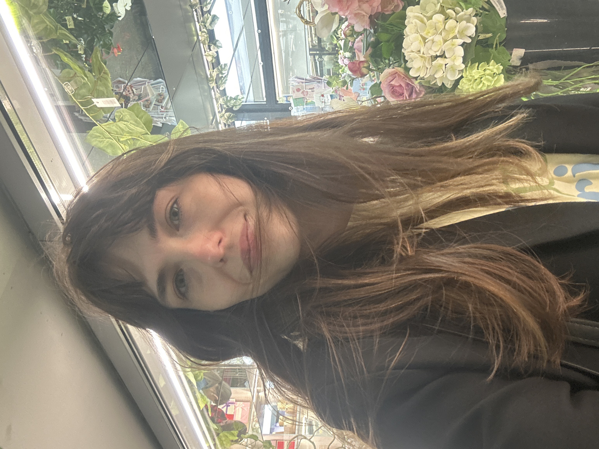 A woman with long, brown hair and bangs is taking a selfie indoors. She is wearing a black jacket over a light-colored shirt with a blue and yellow pattern. Behind her, artificial plants and flowers are displayed. The scene appears to be a store or flower shop, possibly located in Saint Petersburg, Russia, given the context provided. The lighting suggests it might be daytime, with fluorescent lights illuminating the area.