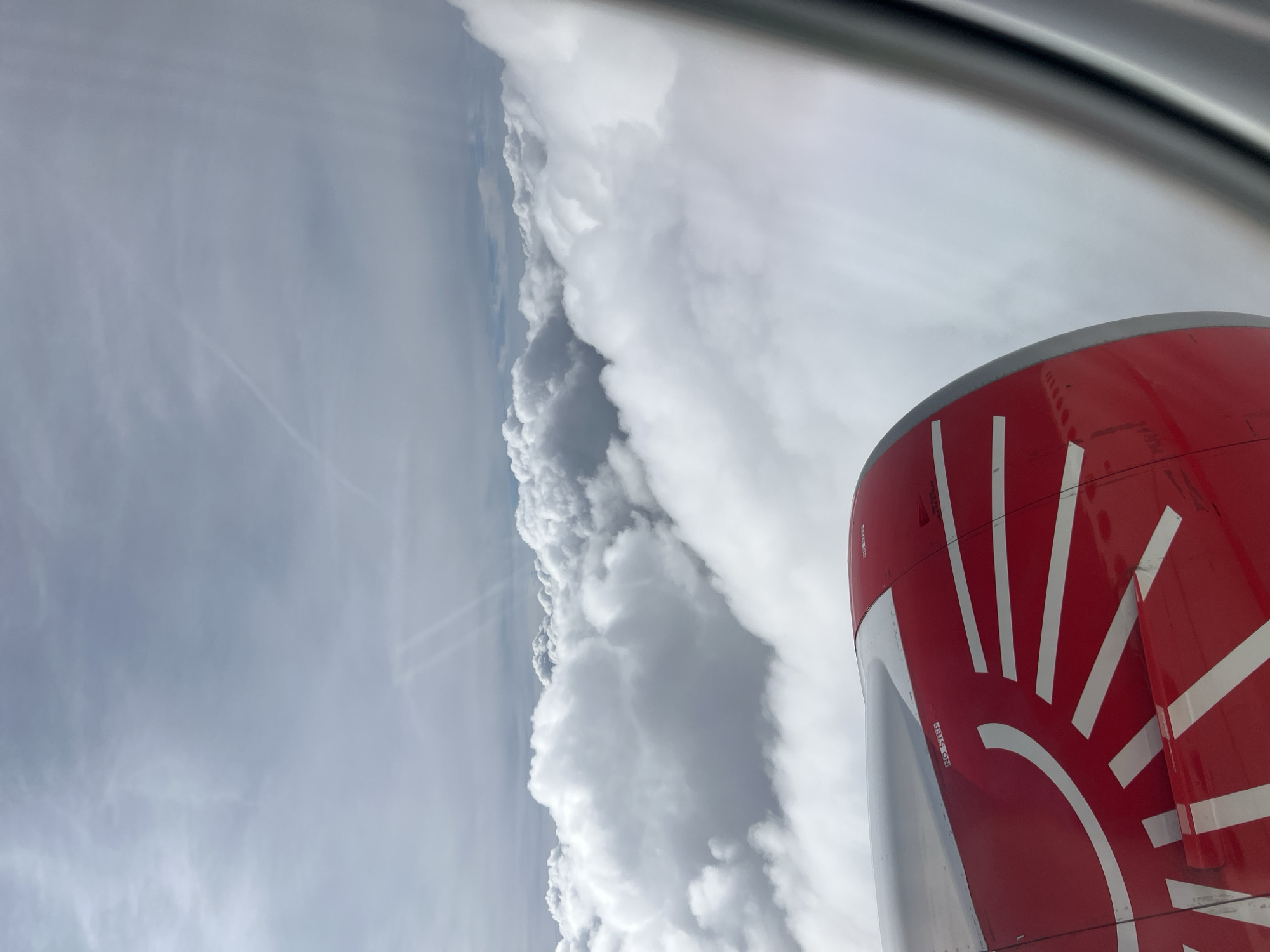 A view from an airplane window reveals a red engine with a white sunburst design. Outside the window, a wall of clouds stretches along the horizon. Above, the sky is a light gray. No city is visible.