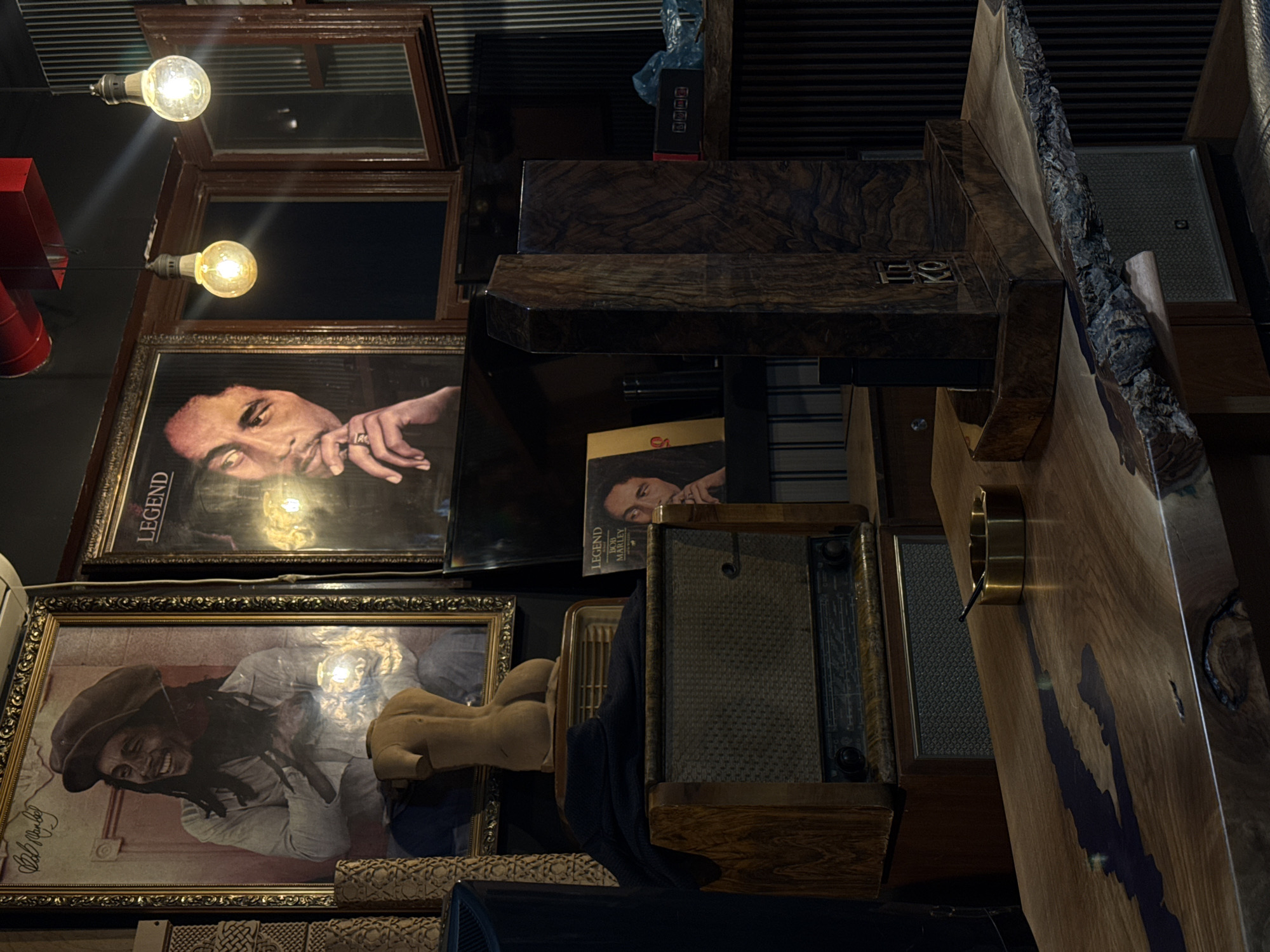 The image shows a cluttered interior space with various items stacked and displayed in a seemingly haphazard manner. Two framed portraits of Bob Marley are prominent, along with a "Legend" album cover. A vintage radio, wooden furniture pieces with unique designs, and other decorative objects are also visible. The lighting is dim, with exposed light bulbs adding to the ambiance. The scene gives the impression of a bohemian or eclectic setting, possibly a bar or lounge with a musical theme. The image was taken inside of Calumet Ethnic Lounge Bar in Yerevan, Armenia.