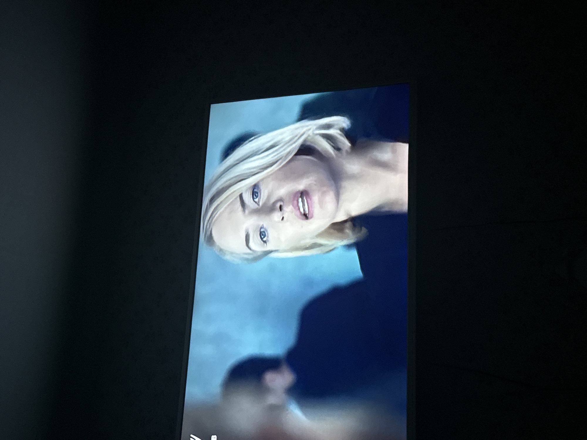 A screen displays a woman with blonde hair and blue eyes. The woman's expression is one of concern or surprise. The background appears to be a blurred image of water or a similar aquatic environment. The screen is surrounded by a dark, nondescript setting, possibly a room. The image's location in the city of Mytishchi, Russia, cannot be determined from the visual information provided.