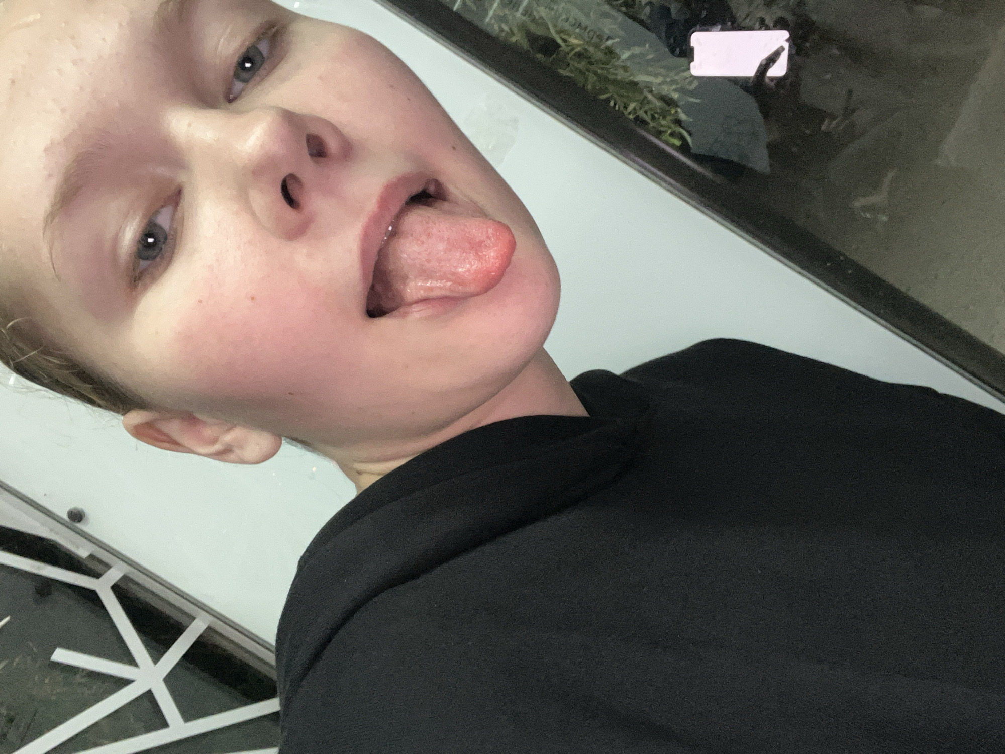 The image is a close-up, indoor selfie of a young Caucasian person with blonde hair and fair skin. They are wearing a black hoodie and sticking out their tongue. The background appears to be a white wall with some geometric patterns on the left side and a dark reflective surface on the right. The lighting suggests an artificial indoor environment. There is no discernible indication of the location being in the city of Perm, Russia.