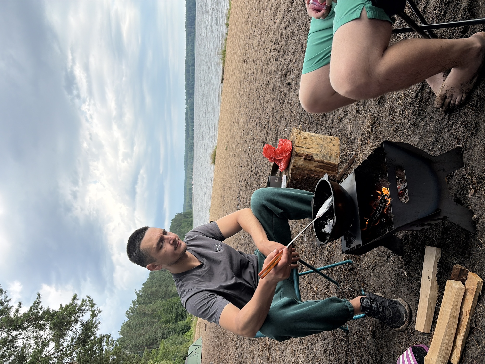 The image depicts an outdoor scene on a sandy beach beside a body of water, likely a lake, with a forest in the background. The sky is overcast with scattered clouds. The setting is Vaskelovo, Russia.
In the foreground, a man is seated on a blue folding chair. He is wearing a dark grey t-shirt and dark green sweatpants. He is holding a cooking utensil with an orange handle and appears to be stirring something in a black pot placed on a portable black grill. The grill has visible flames and burning wood. Beside the grill is a tree stump with a red plastic bag and what looks like a metal container on it. To the left of the man, there are pieces of firewood.
To the right of the frame, another person's legs are visible. They are wearing bright green shorts and are seated on a chair, part of which is visible. This person's bare feet are covered in mud.
The overall impression is one of a camping or outdoor cooking scenario.