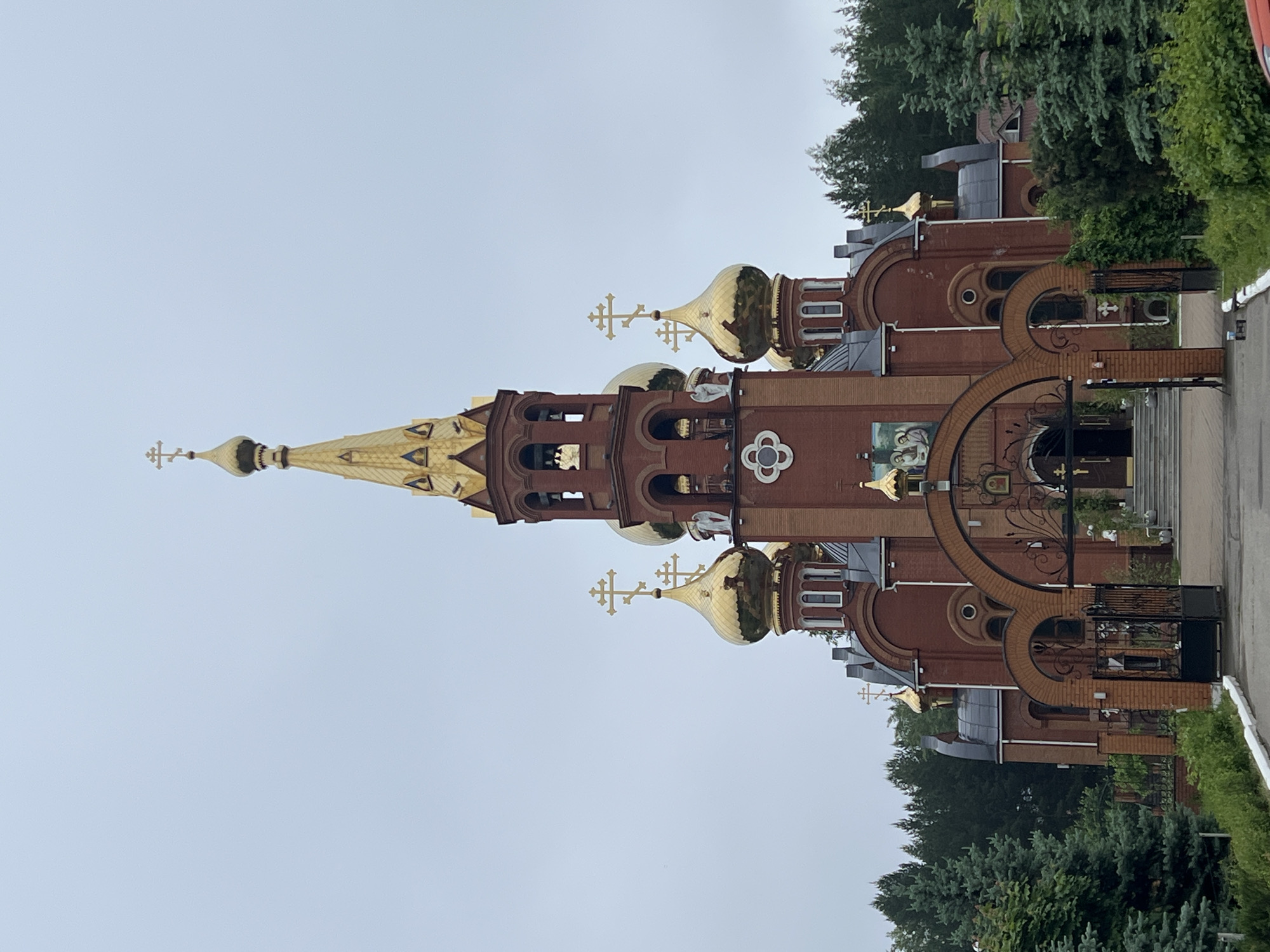 This image captures an ornate Eastern Orthodox church located in Chaikovsky, Russia, under an overcast sky. The church is predominantly constructed from reddish-brown brick and is characterized by multiple golden onion domes and a tall, slender central spire, each crowned with a distinctive Orthodox cross.
The architectural style includes numerous arched windows and doorways, intricate brick detailing, and decorative elements. A prominent white, four-lobed motif adorns the main facade, below which a large painted icon depicting religious figures is integrated into the brickwork. To the right, an elaborate dark metal gate with decorative scrolls and small crosses provides access to the church grounds, set within a grand brick archway.
Lush green deciduous and coniferous trees frame the church, with dense foliage visible in the background and foreground, particularly on the right side. A paved path or driveway runs along the right edge of the scene. The sky is a uniform light grey, indicating an overcast daytime. No people or specific activities are visible. While there are decorative elements on the gate and the icon, no legible text is discernible.