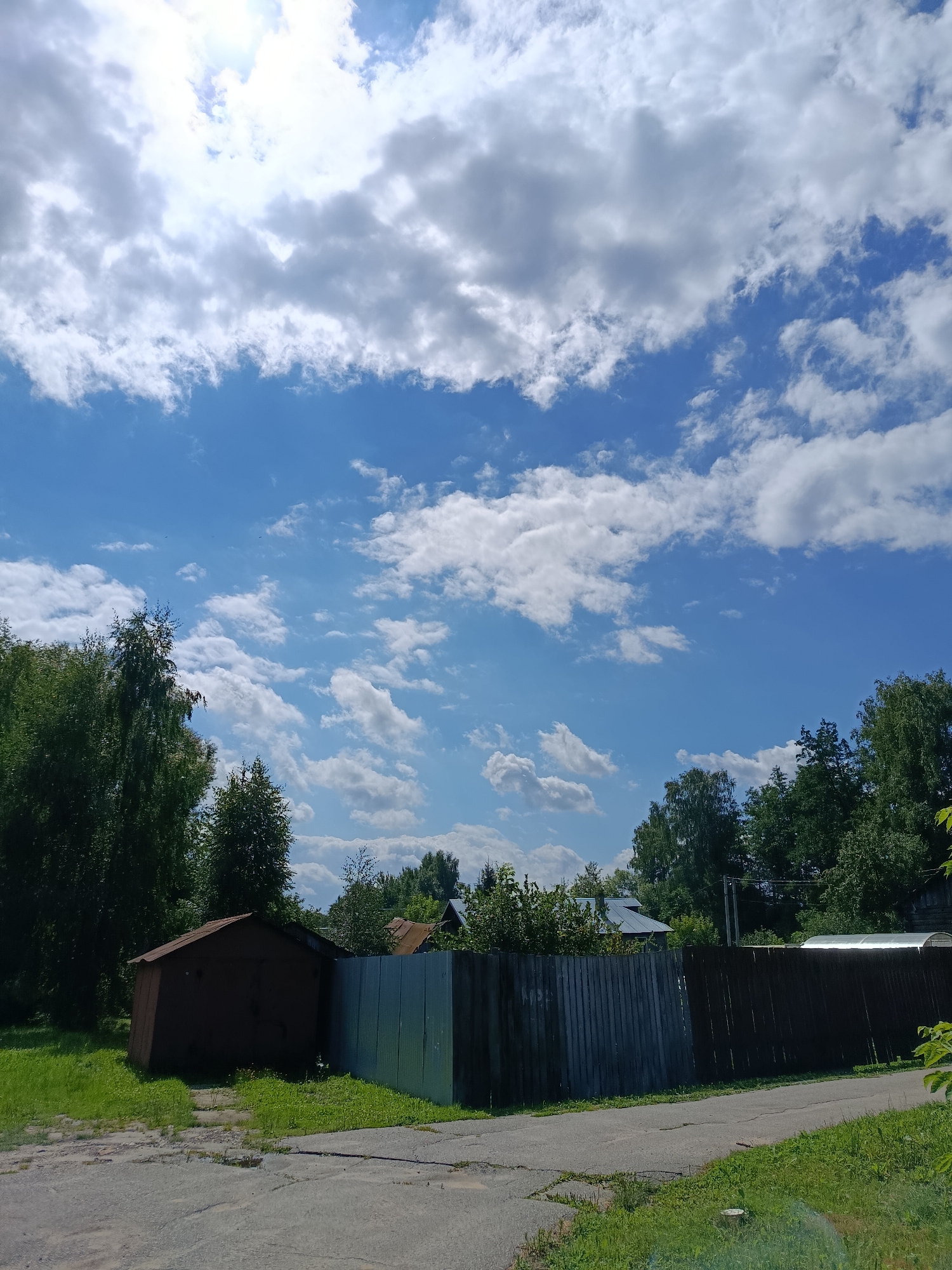 The image depicts a rural scene in Vareya, Russia, under a bright blue sky filled with scattered white clouds. In the foreground, a paved road or driveway cuts through a grassy area. To the left of the road is a dark brown, single-story building with a metal roof, possibly a garage or shed, situated on a patch of grass. Adjacent to this structure is a light gray corrugated metal fence. Further to the right, a tall, dark wooden fence runs horizontally across the frame. Behind the fences and buildings, a dense line of green trees forms a natural barrier. In the distance, glimpses of rooftops and more trees are visible, suggesting a residential area or village. The overall atmosphere is one of a sunny, perhaps summer, day in a tranquil, non-urban setting.