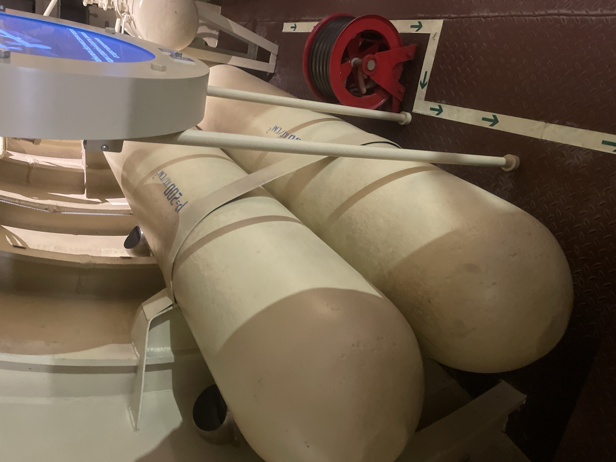 The image shows two large, cylindrical, cream-colored objects, possibly torpedoes or ballast tanks, lying horizontally. They are secured by beige straps. One object has blue text that appears to read "P-200 KICM²". The objects are in an indoor environment with cream-colored, curved walls and metallic structures. To the right, a red reel with a dark hose is visible on the floor. White and green directional arrows are marked on the dark floor. In the background, a blurred screen displays blue text and graphics. This scene is located in the Museum of Naval Glory in Kronstadt, Russia, part of the "Island of Forts" museum-defense complex.