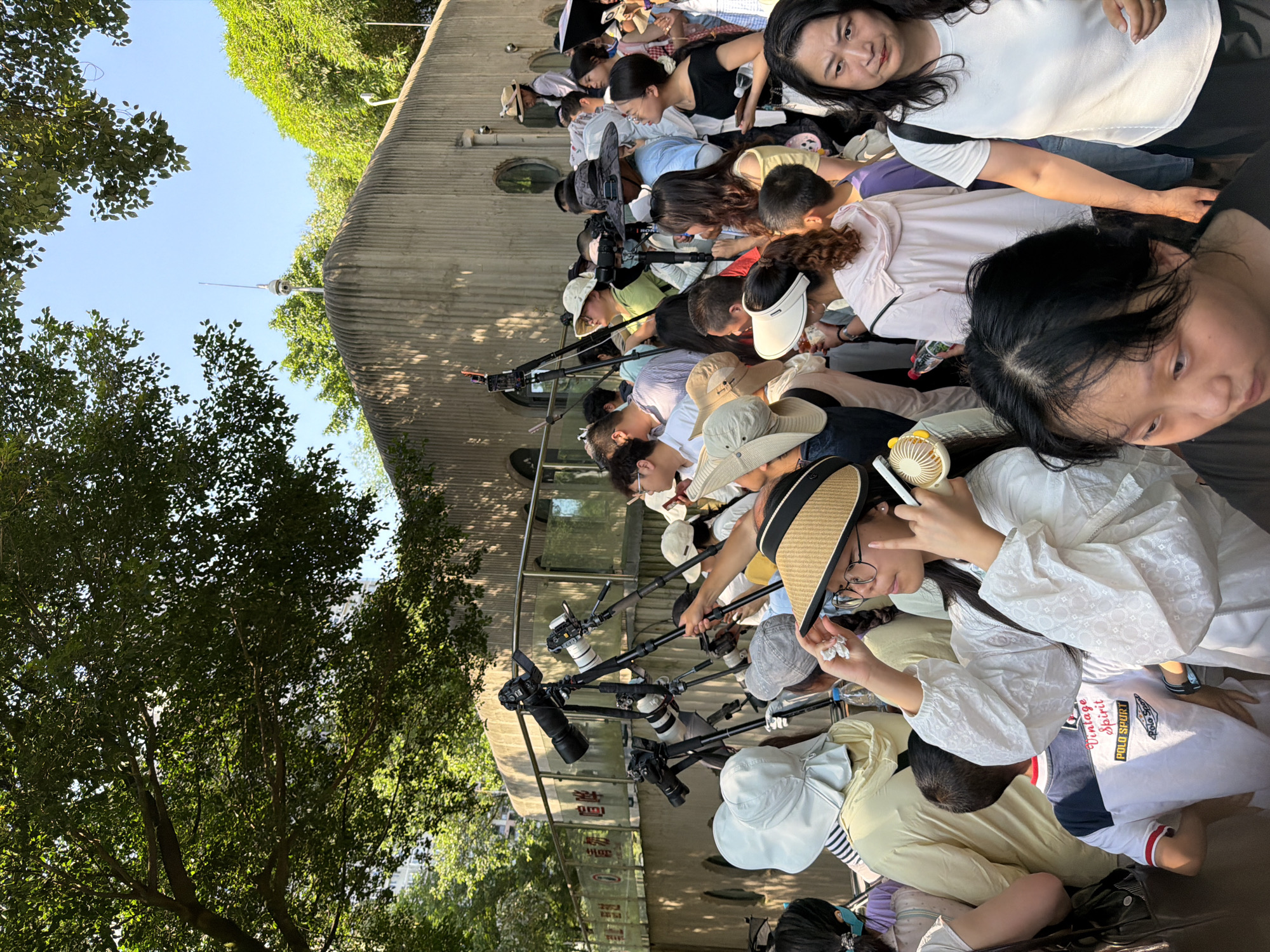 In the Jiulongpo District of China, a crowd of people are gathered outdoors under a tree-lined sky. Many individuals are holding cameras on tripods, suggesting they are photographers or enthusiasts observing an event. The background features a concrete building with a ribbed texture, and on its roof, a security camera is visible atop a pole. The scene appears to be a public space, possibly a park or an area with an attraction, given the presence of both onlookers and professional-looking camera equipment. The light suggests it is daytime, and the presence of a fan on one person's head indicates warm weather.