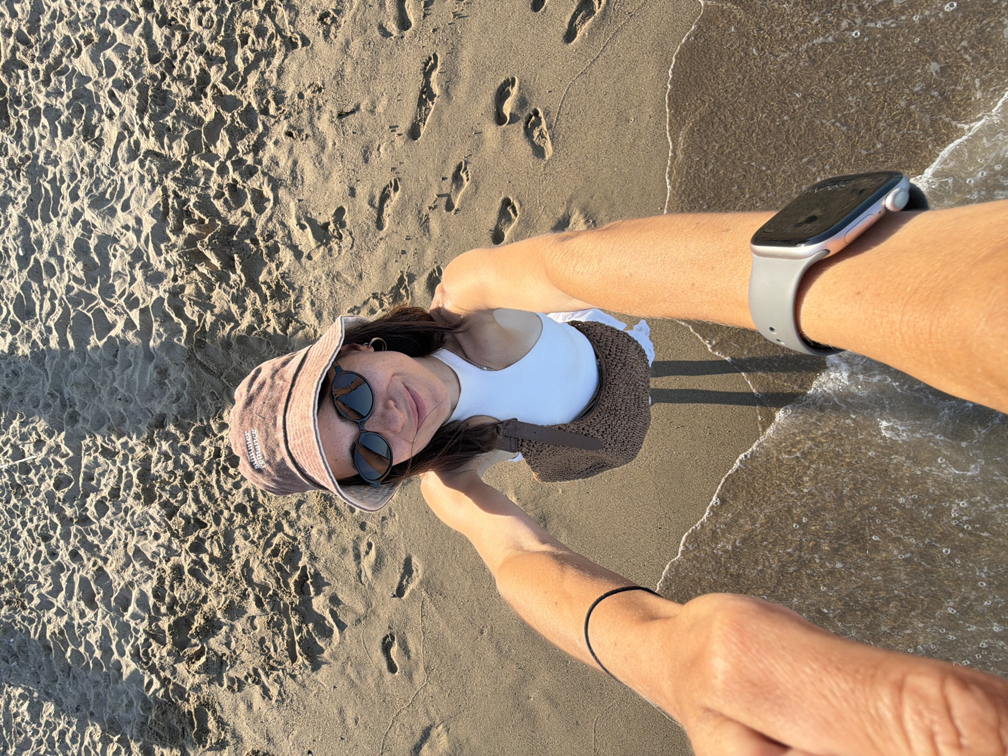 A selfie taken from a high angle shows a young woman standing on a sandy beach, with the edge of the water in the foreground. She is wearing a white tank top, a light brown bucket hat, and sunglasses. A woven brown bag is slung over her shoulder. Her left arm is extended towards the camera, and on her wrist, she is wearing a gray smartwatch. The sand is textured with footprints and shadows cast by an unseen object, possibly an umbrella. The scene is set on a beach, but there is no specific information to suggest it is in Barcelona, Spain.