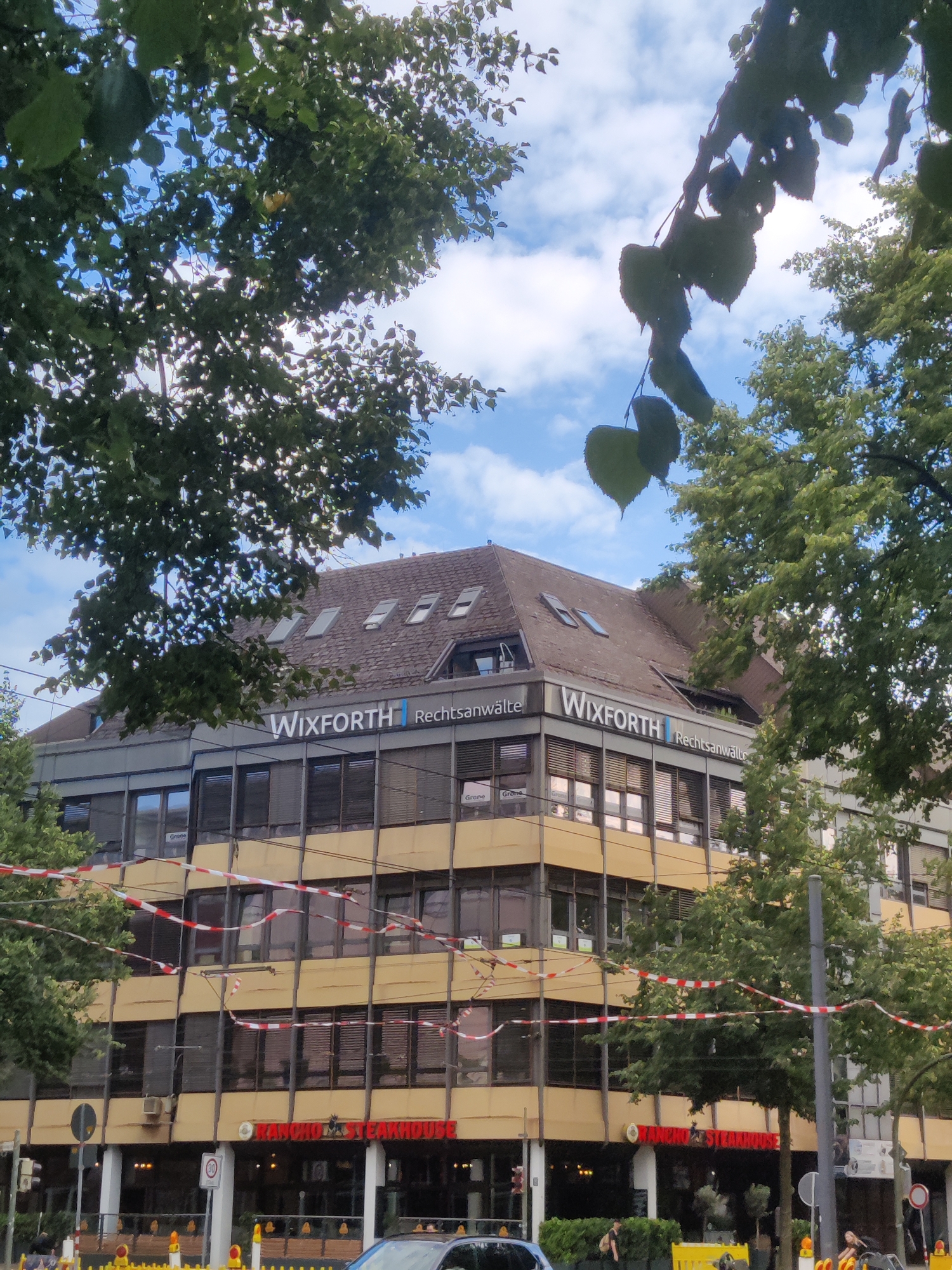 The image shows a building in Bielefeld, Germany, with the signage "WIXFORTH Rechtsanwälte" visible on the upper floors, indicating a law firm. Below this, the text "RANCHO STEAKHOUSE" is prominently displayed, identifying a restaurant on the ground level. The building is modern, with multiple floors featuring numerous windows and a distinctive yellow facade. Red and white striped tape is strung across the facade, possibly indicating construction or a restricted area. Trees with green foliage partially obscure the building, and the sky above is visible with scattered clouds. Several streetlights and traffic signals are present, suggesting an urban setting. The general scene depicts a street view of a commercial building housing both a professional service and a dining establishment.