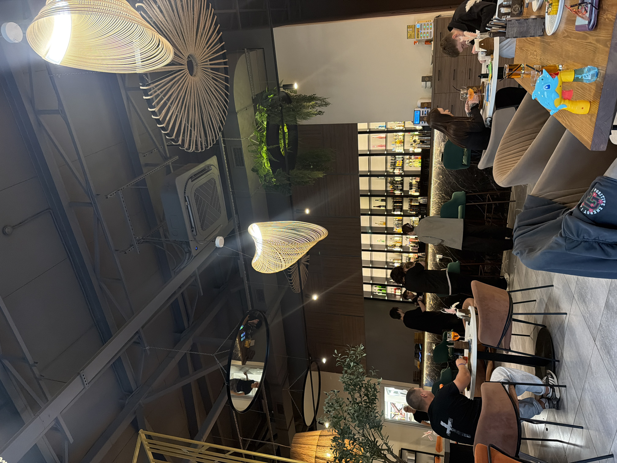 The image depicts the interior of a modern restaurant or bar, possibly located in an urban setting. The scene is illuminated by ambient lighting, with several decorative light fixtures hanging from the ceiling, including a prominent spiral-shaped one and a circular, fan-like fixture. A bar area is visible in the background, stocked with various bottles of liquor and illuminated by spotlights.
In the foreground, several patrons are seated at tables. One man, wearing a black t-shirt with a white cross graphic and light-colored pants, is seated at a round table with a drink and appears to be eating. Another table to the right has two individuals, a woman with long dark hair and a man, both engaged with their meals or drinks. In the middle ground, several staff members are visible behind the bar, attending to their duties.
The restaurant's decor features a mix of materials, including dark wood paneling behind the bar and on the walls, and sleek, modern furniture, primarily in muted tones of brown and gray. There are also some decorative plants, including a potted olive tree on the left side of the frame. The flooring is a light-colored tile with a subtle geometric pattern. The overall atmosphere appears to be relaxed and contemporary.
