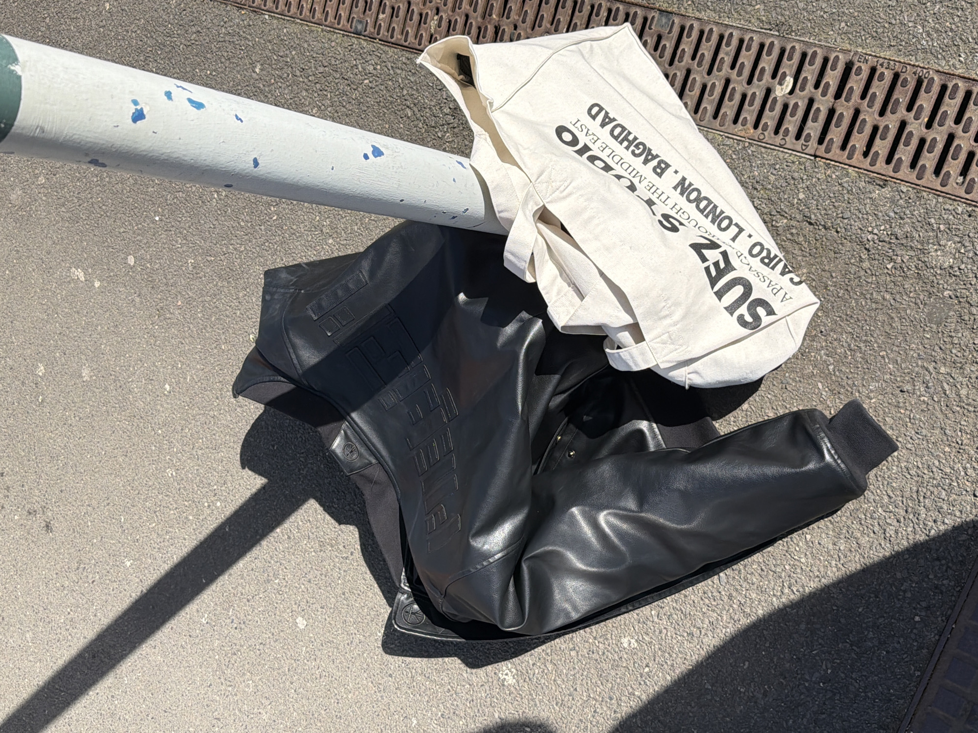 In London, United Kingdom, a black faux leather jacket and a cream canvas tote bag are positioned on gray asphalt ground. The jacket, featuring ribbed cuffs and faintly embossed text on its back, is partially folded. Adjacent to the jacket, the tote bag displays the black text "SUEZ STUDIO / A PASSAGE THROUGH THE MIDDLE EAST / CAIRO. LONDON. BAGHDAD". Both items rest near a white pole, which has a green cap and shows blue paint peeling from its surface. A rectangular metal drainage grate, marked "EN 1433 D 400", runs parallel to the pole and bag on the right side of the frame. Strong sunlight casts distinct shadows across the scene. No individuals are present.