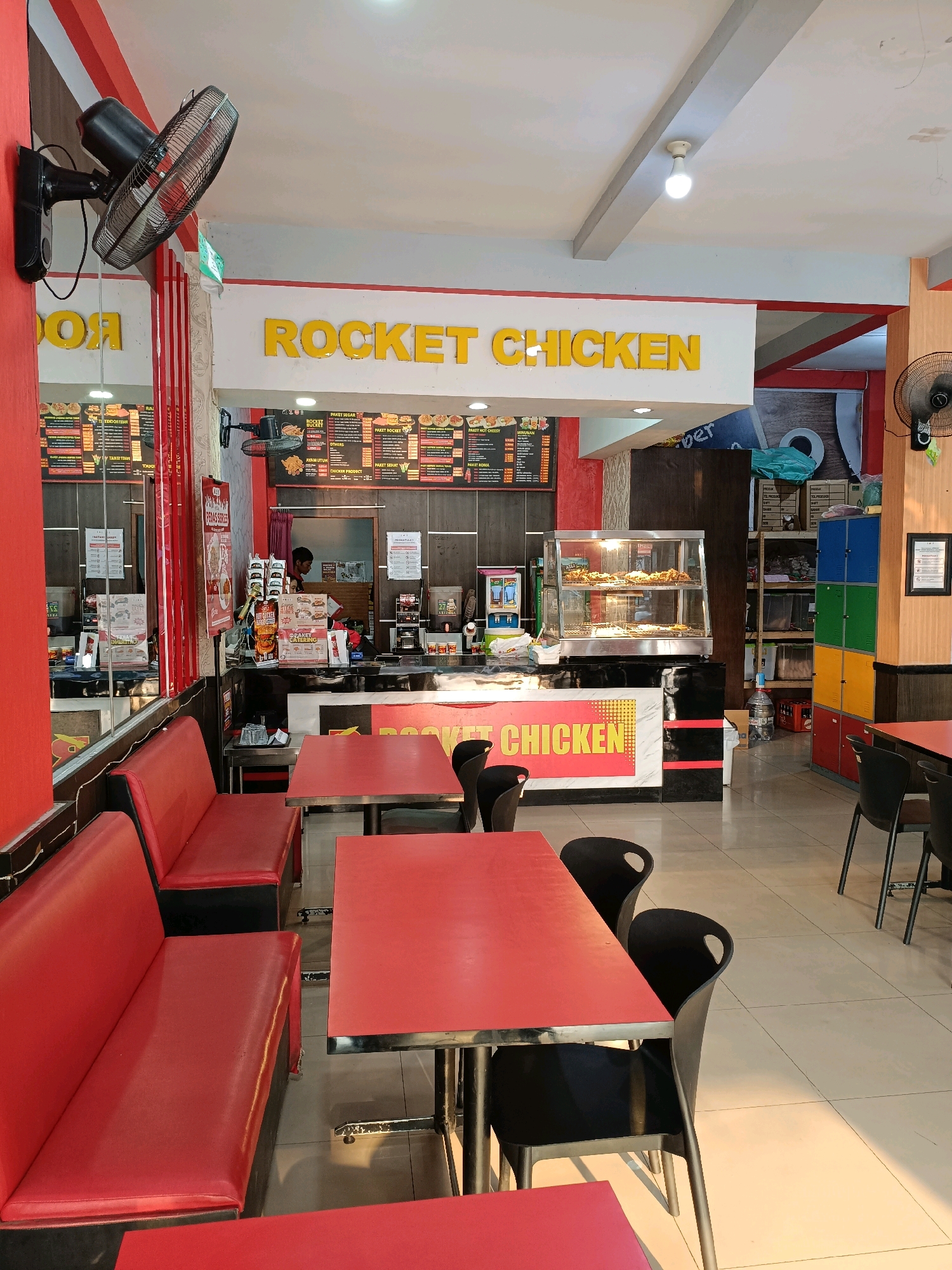 The image depicts the interior of a fast-food restaurant called "Rocket Chicken" in Ngemplak, Indonesia. The restaurant features red booth seating with black accents, accompanied by red tabletops and black chairs. The walls are painted in red and white, with the restaurant's name prominently displayed in yellow lettering above a counter area. Behind the counter, a glass display case showcases fried chicken. A person is visible behind the counter, potentially an employee. The environment includes various restaurant equipment, menus displayed on the walls, and storage shelves in the background. The flooring appears to be tiled.