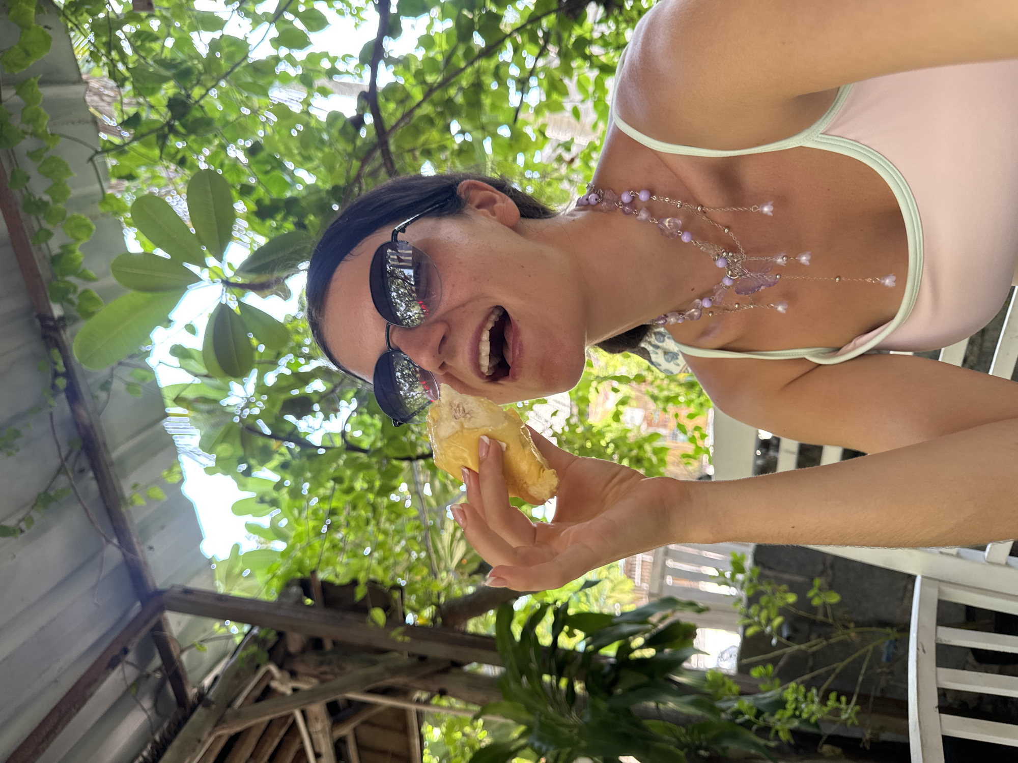 A young woman with dark hair and sunglasses is taking a bite of durian fruit while sitting outdoors in Mũi Né, Vietnam. She is wearing a light pink tank top with a pale green trim and a long, layered necklace with purple beads and charms. Her left hand holds the durian to her mouth. The background shows lush green foliage and a rustic, open-air roof structure made of metal and wood. She appears to be seated on a white chair.