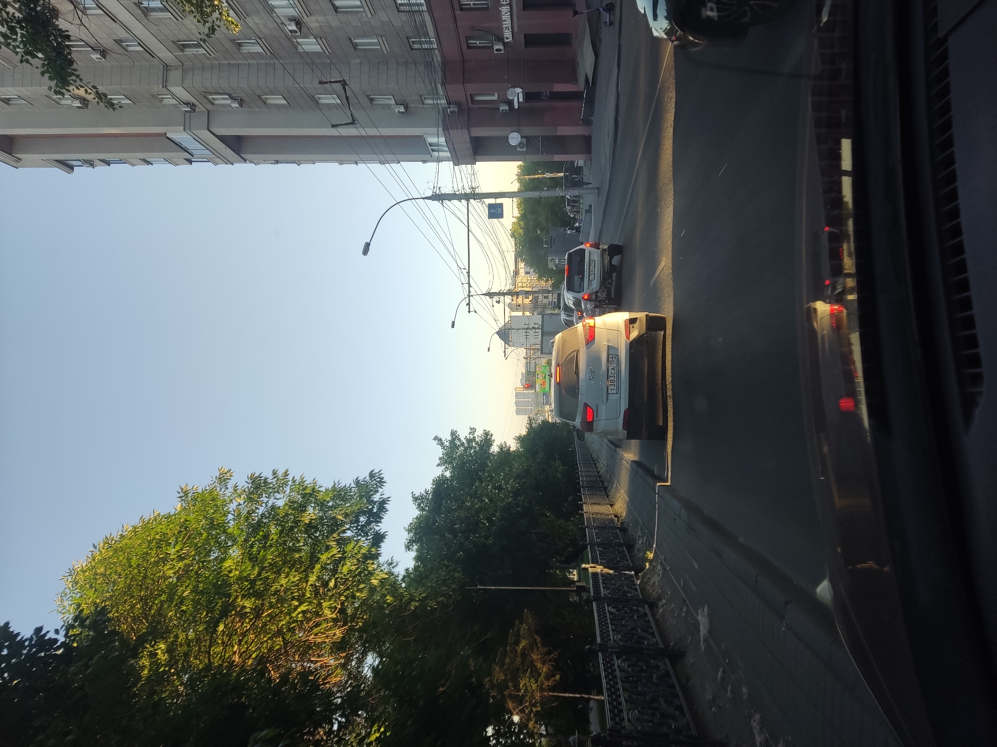 The image shows a street scene in Novosibirsk, Russia, on a clear day with abundant sunlight casting long shadows. The perspective is from inside a vehicle, looking forward. On the left side of the street, there is a sidewalk lined with an ornate black metal fence, followed by a row of lush green trees casting dappled shade. Beyond the trees, more distant buildings and greenery are visible.
The road in front is occupied by several vehicles, suggesting moderate traffic. The most prominent car in the foreground is a white SUV, likely a Hyundai Creta based on the visible badging, traveling in the same direction. Another SUV, a dark color, is in the lane to the right. Further ahead, a light-colored sedan is visible, also in the same lane as the white SUV. Traffic lights and street lamps are present.
To the right of the road, a multi-story building with a light gray facade and darker reddish-brown lower levels dominates the skyline. The building features numerous windows and air conditioning units. Signs are visible on the lower part of the building, one of which appears to read "Хильдсбой" in Cyrillic. A scooter is parked on the sidewalk in front of this building.
The sky is a clear, light blue, indicating fair weather. The overall impression is an urban environment captured during the golden hour, with sunlight illuminating the scene and creating a warm atmosphere.
