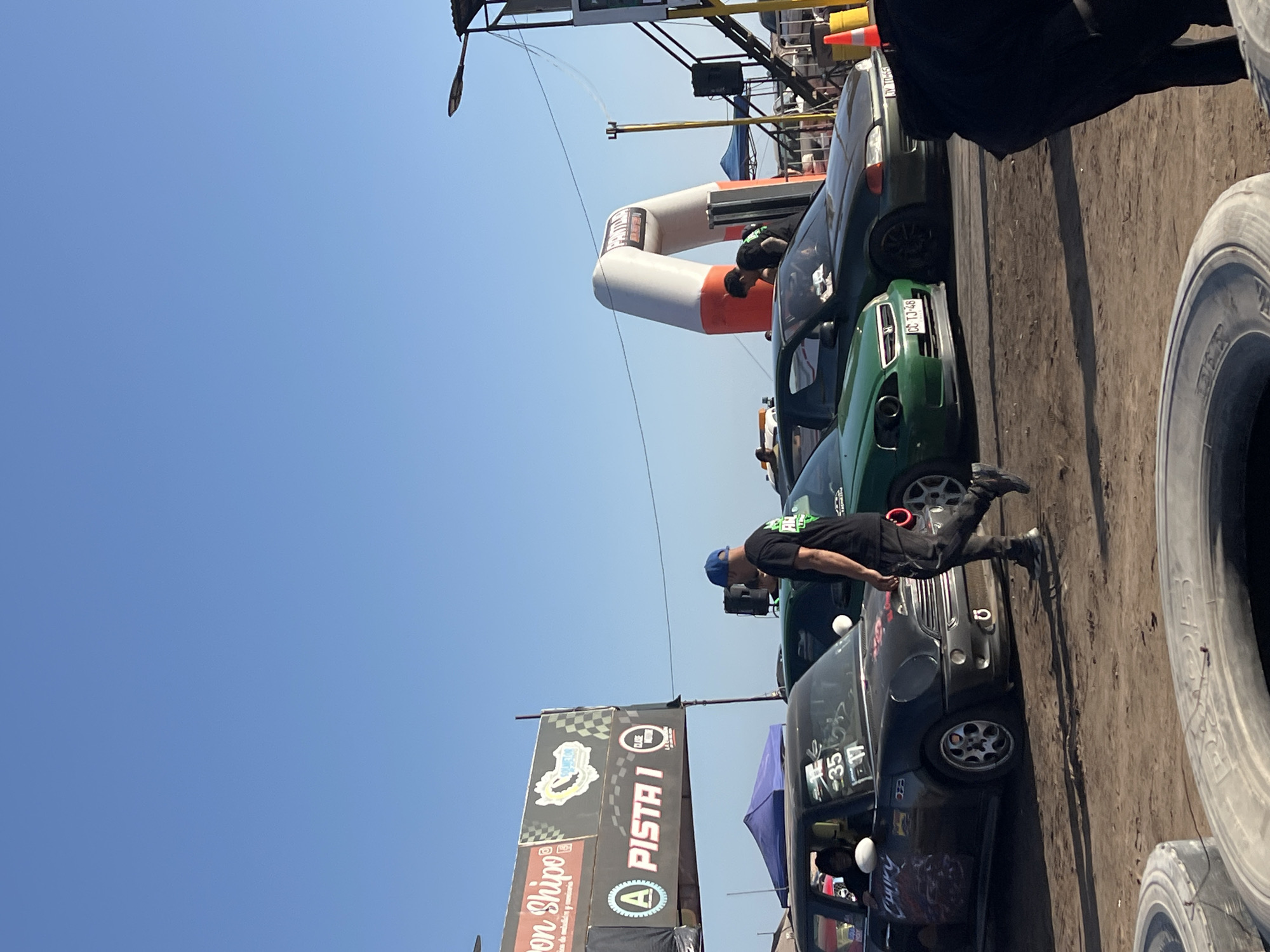 The image shows a daytime outdoor scene, presumably at a racing event or car meet. A line of cars is parked close to a dirt or gravel ground surface. Several individuals are seen around the vehicles. One man is bending down next to a dark-colored car, possibly examining it or talking to someone inside. A large inflatable arch with orange and white stripes is visible, indicating a start or finish line. A banner with "A PISTA 1" is displayed, along with other logos and text. Tires are stacked next to the cars. The scene is likely set in the city of Arica, Chile based on the OCR results.