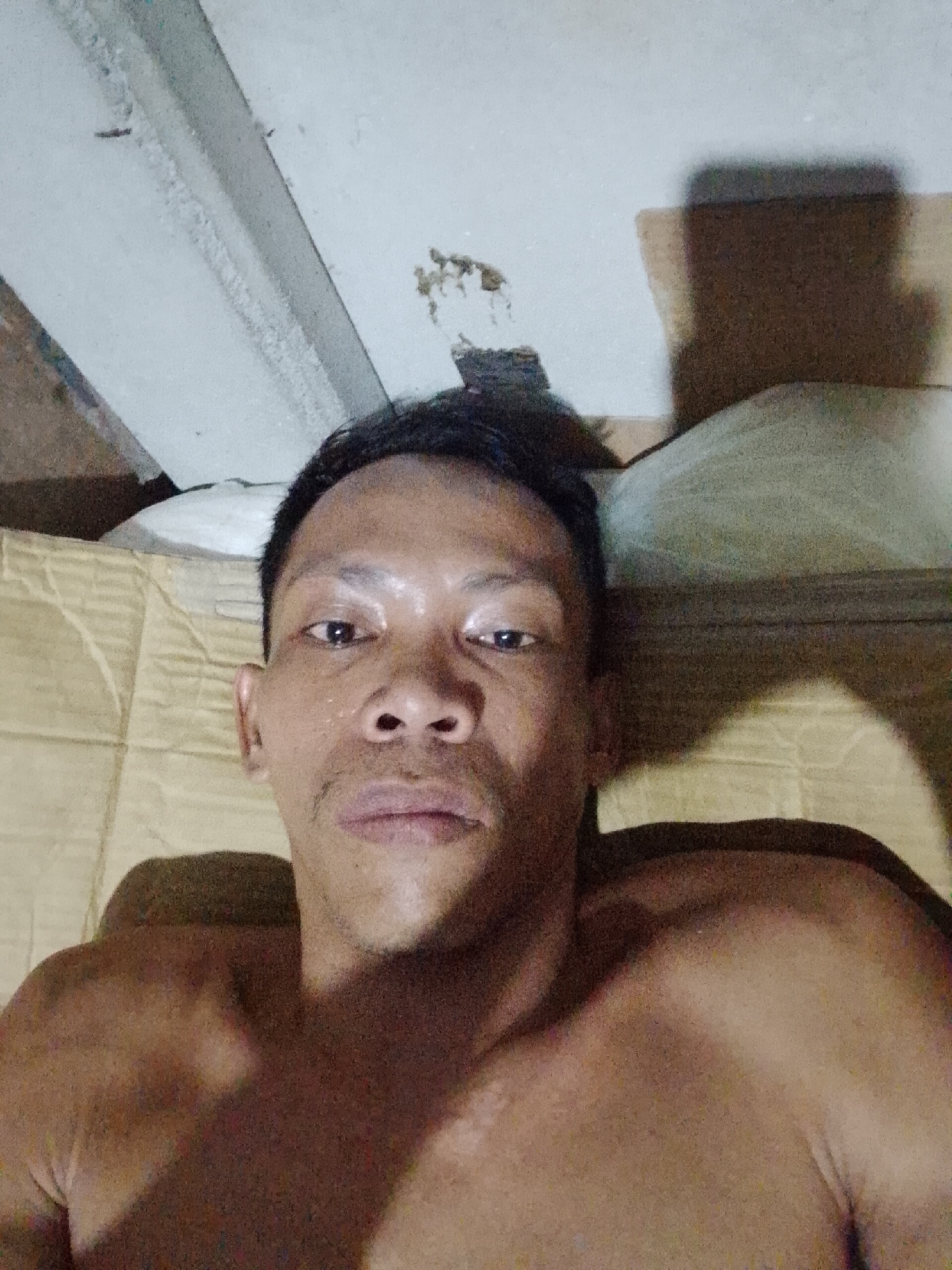 The image is a selfie of a shirtless, Southeast Asian man with short, dark hair. He is lying down on a patterned beige surface, presumably a bed. The background includes a white ceiling with some exposed elements and a shadowy area with what appears to be a pillow or other soft object.
The location is unknown. The mention of Bukit Ketok, Indonesia cannot be verified from the image.