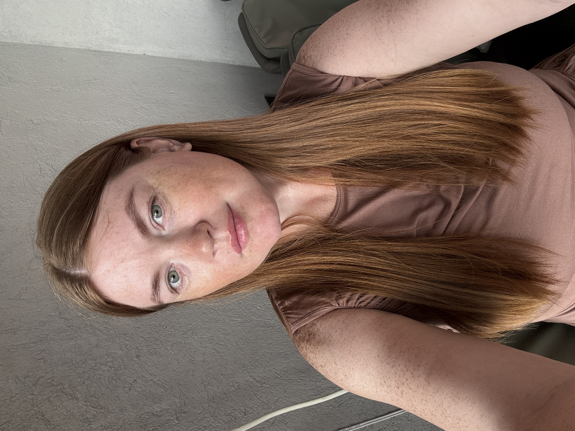 A young woman with fair skin, freckles, and long auburn hair is shown in a close-up, waist-up selfie. She has green eyes and is wearing a brown, sleeveless top. The background appears to be an interior setting with a textured gray wall on the left and a lighter textured wall on the right. A white cable is visible on the left side of the frame. The image does not provide any specific details or context to locate it in the city of Gyumri, Armenia.