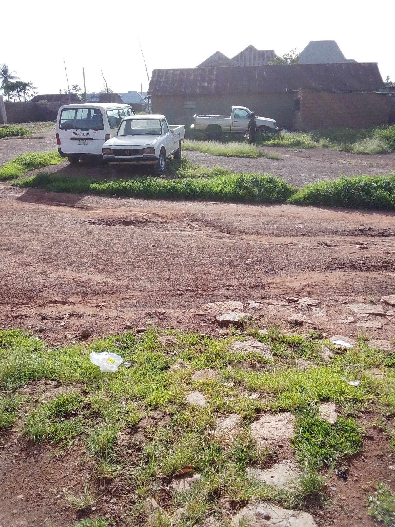 A white van with "EVANGELISM" written on its rear is parked next to a white pickup truck on a dirt road. Behind the vehicles, a long, low building with a corrugated metal roof stands under a bright sky, suggesting it is daytime. The building appears to be made of brick or mud. In the foreground, a patch of overgrown grass is interspersed with stones and dirt. A white plastic bag lies in the grass. To the right of the white pickup truck, a silver pickup truck is parked near the building, and a person is walking away from it, carrying something. The scene is rustic, with evidence of unpaved roads and vegetation, consistent with a rural or semi-rural setting in Mangu, Nigeria.